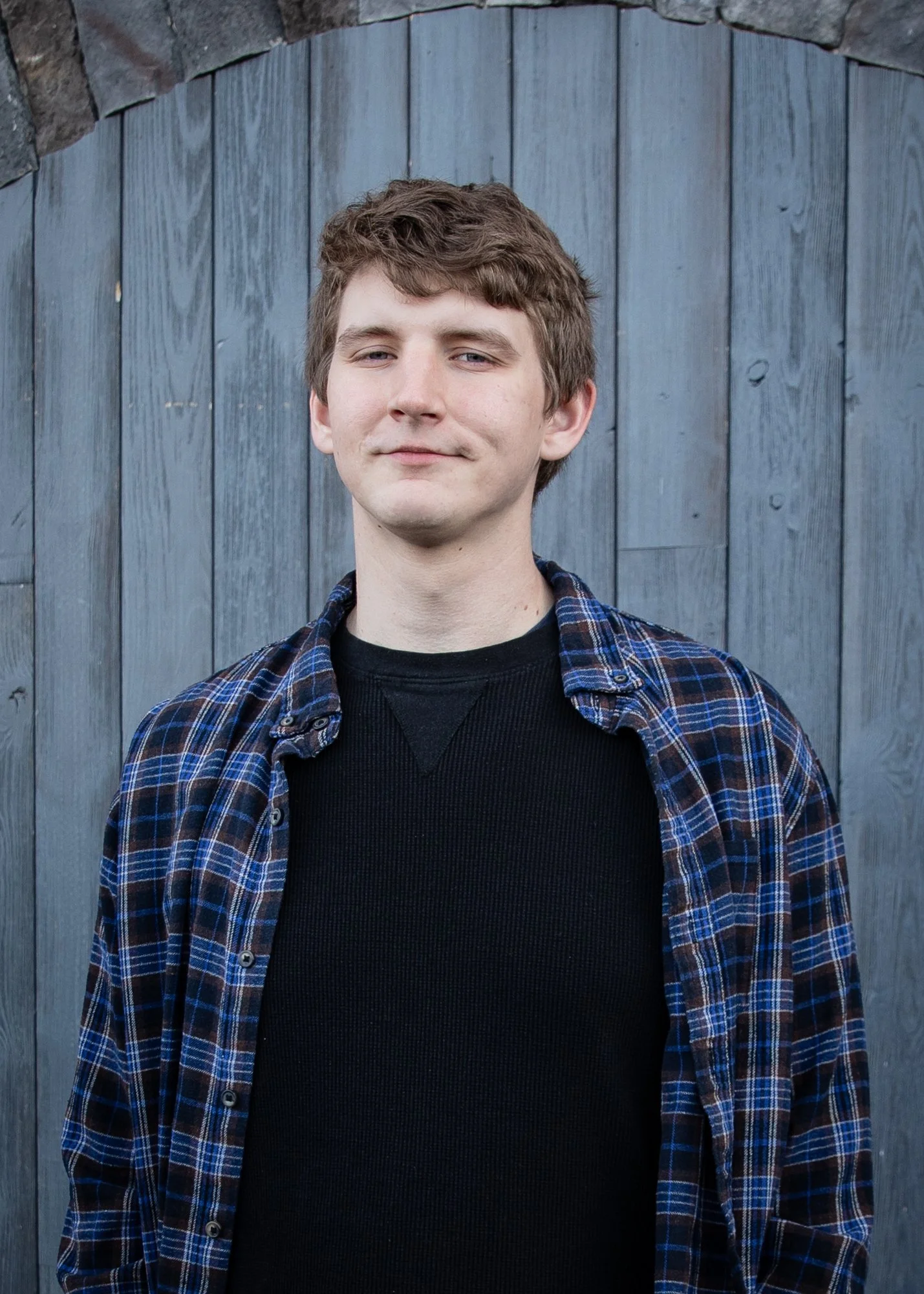 A young man with light skin and wavy brown hair standing in front of a blue wooden fence, wearing a black sweater and a blue plaid shirt.