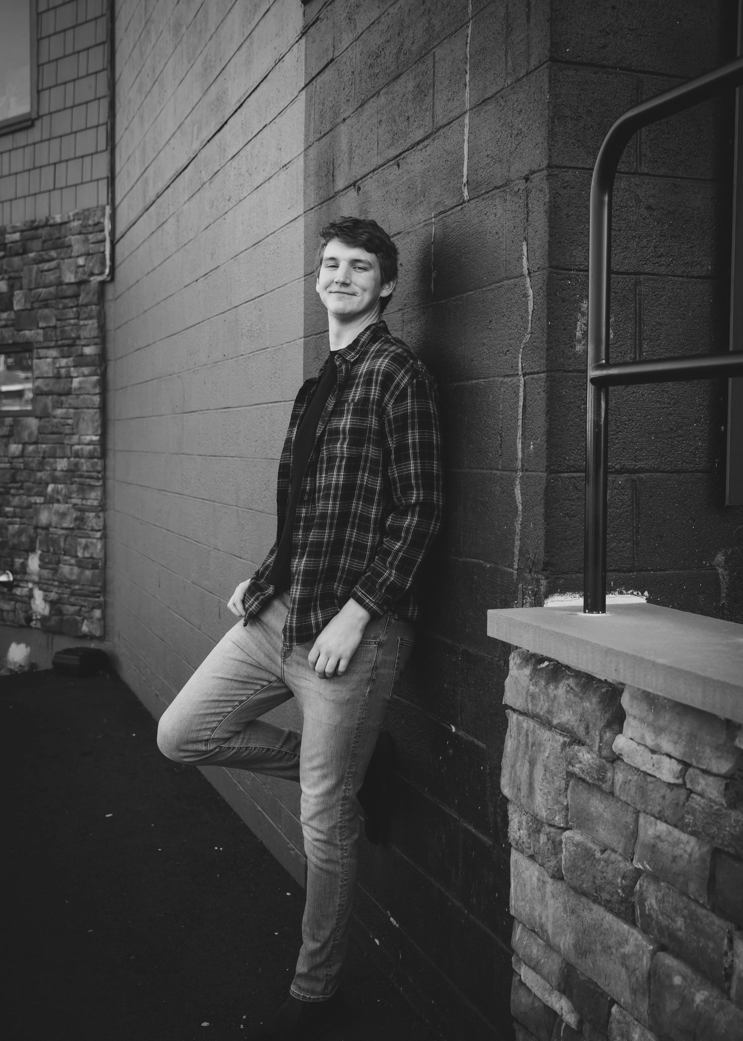 Black-and-white photo of a young man smiling, leaning against a brick and stone wall, with a metal railing nearby.