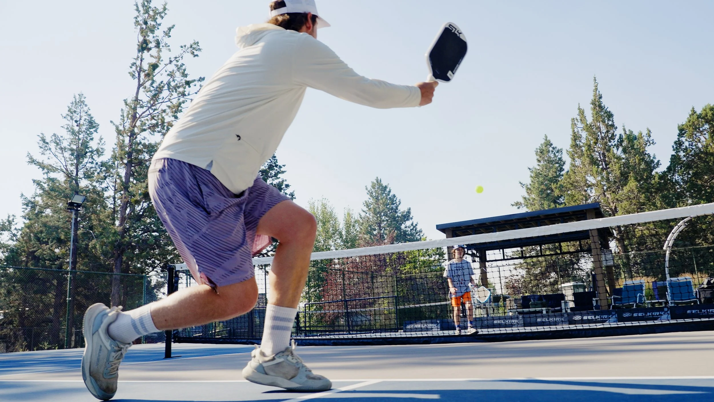 A person is playing pickleball on an outdoor court, swinging a paddle to hit a yellow ball while another person stands in the background near the net.
