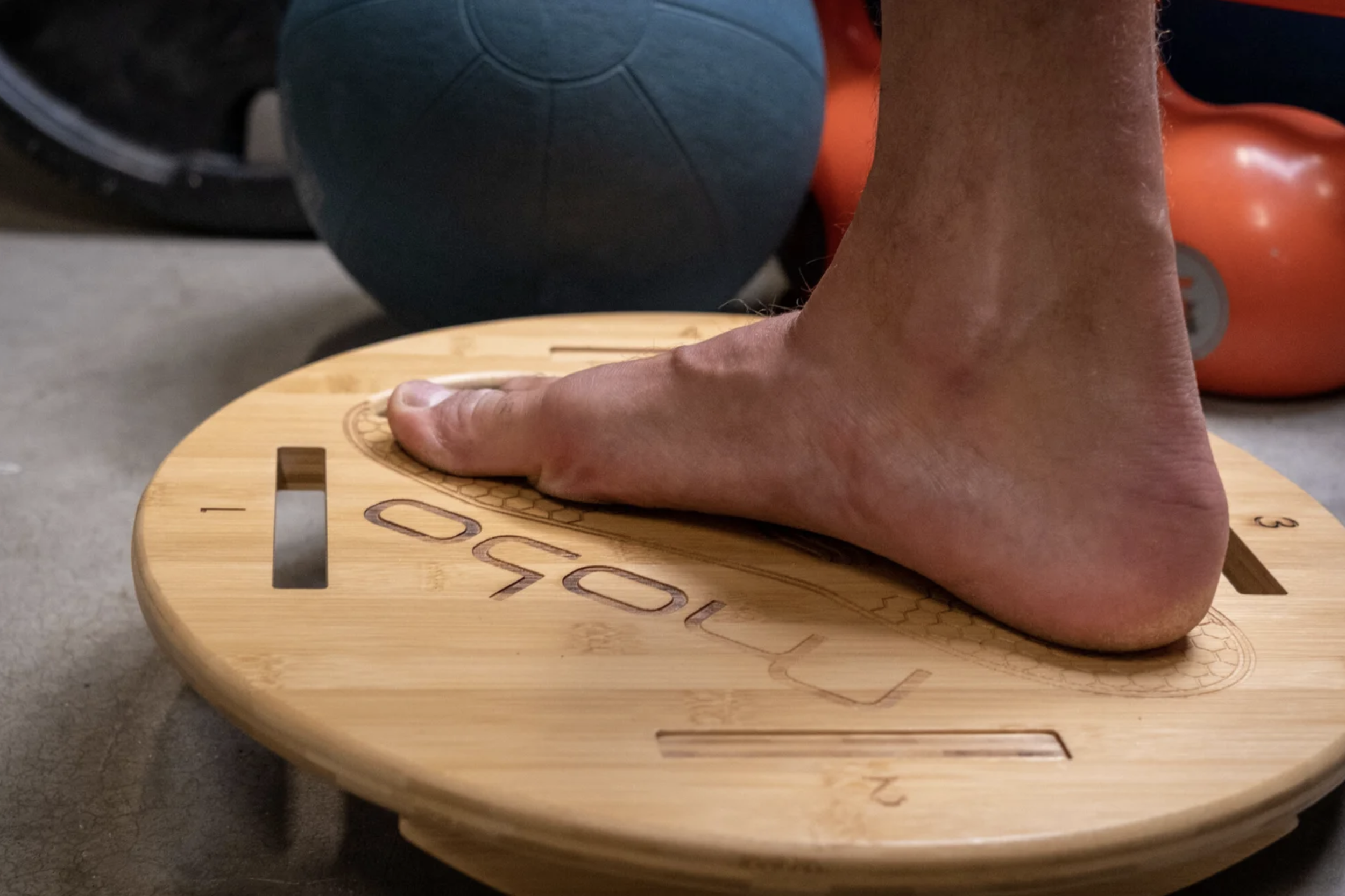 A person performing a handstand with their hand on a wooden balance board, with fitness equipment such as a blue kettlebell and orange weights in the background.