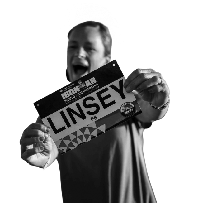 A man smiling and holding a National Ironman World Championship bib with the name 'LINSY' on it.