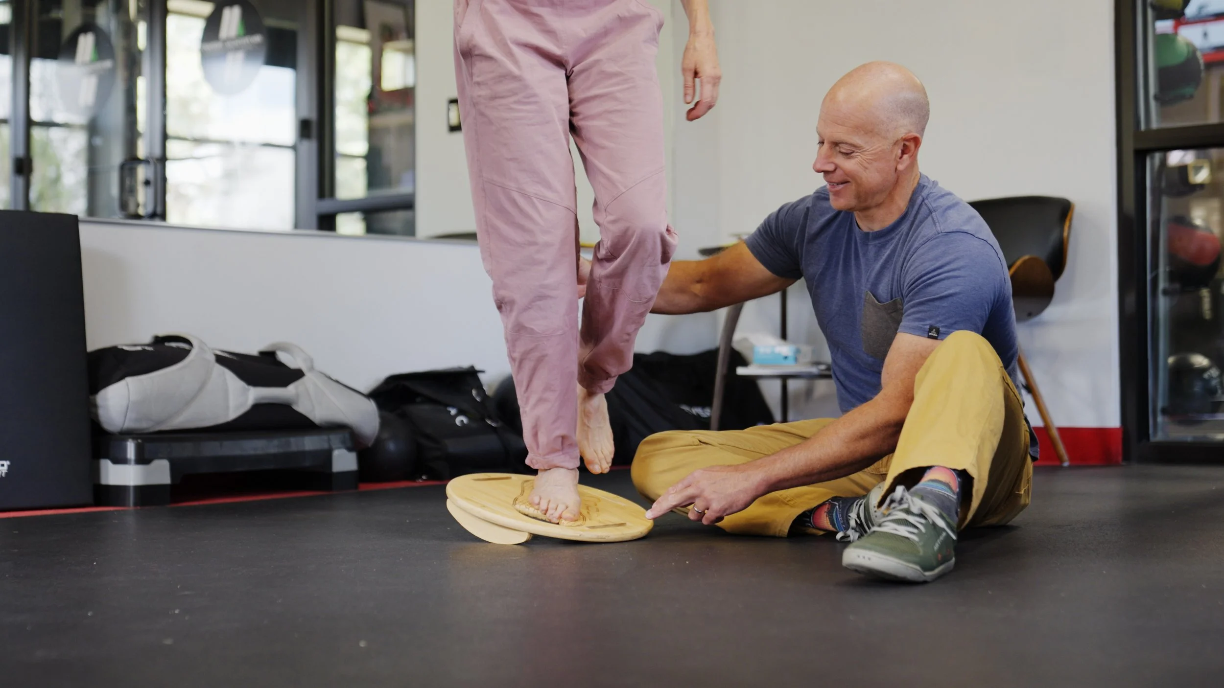 A man helping a woman stand on a balance board in a gym.