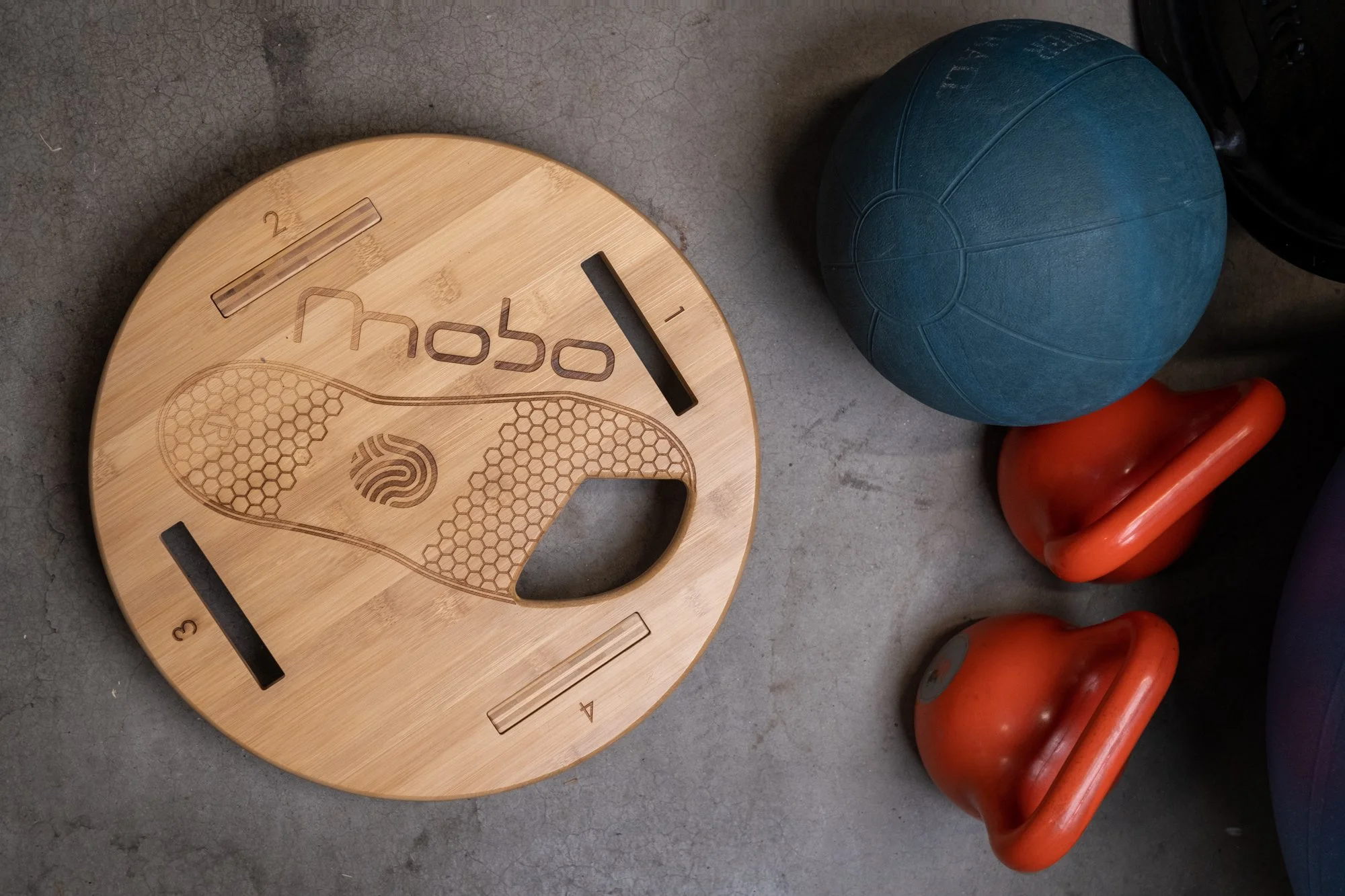 A wooden balance board with the word 'Uplift' engraved on it, a blue medicine ball, and two orange kettlebells on a gray concrete floor.