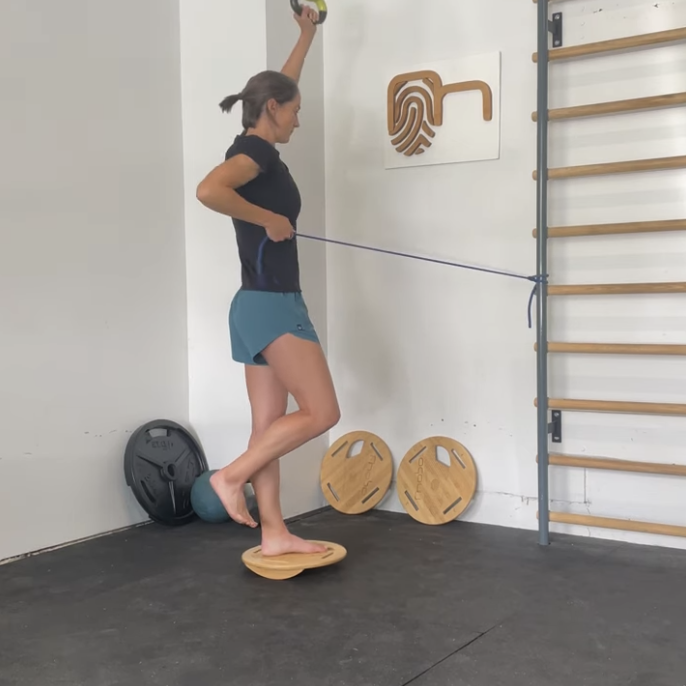 A woman balancing on a wooden wobble board while performing a physical exercise with a resistance band, in a gym setting with workout equipment and wall art.