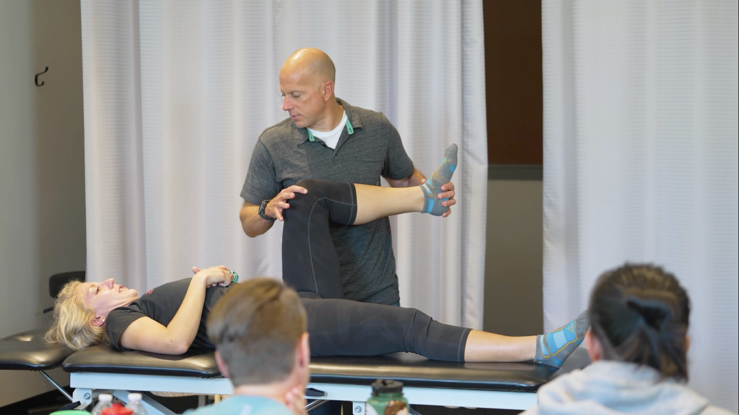 A physical therapist demonstrates a leg exercise to a woman lying on a treatment table, with other people observing in a clinical setting.