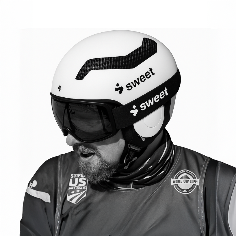A man wearing a white racing helmet with black carbon fiber accents, black goggles, and a black neck gaiter, dressed in a dark jacket with various patches and logos, including 'STIF US ART TEAM' and 'WORLD CUP SUSP'.