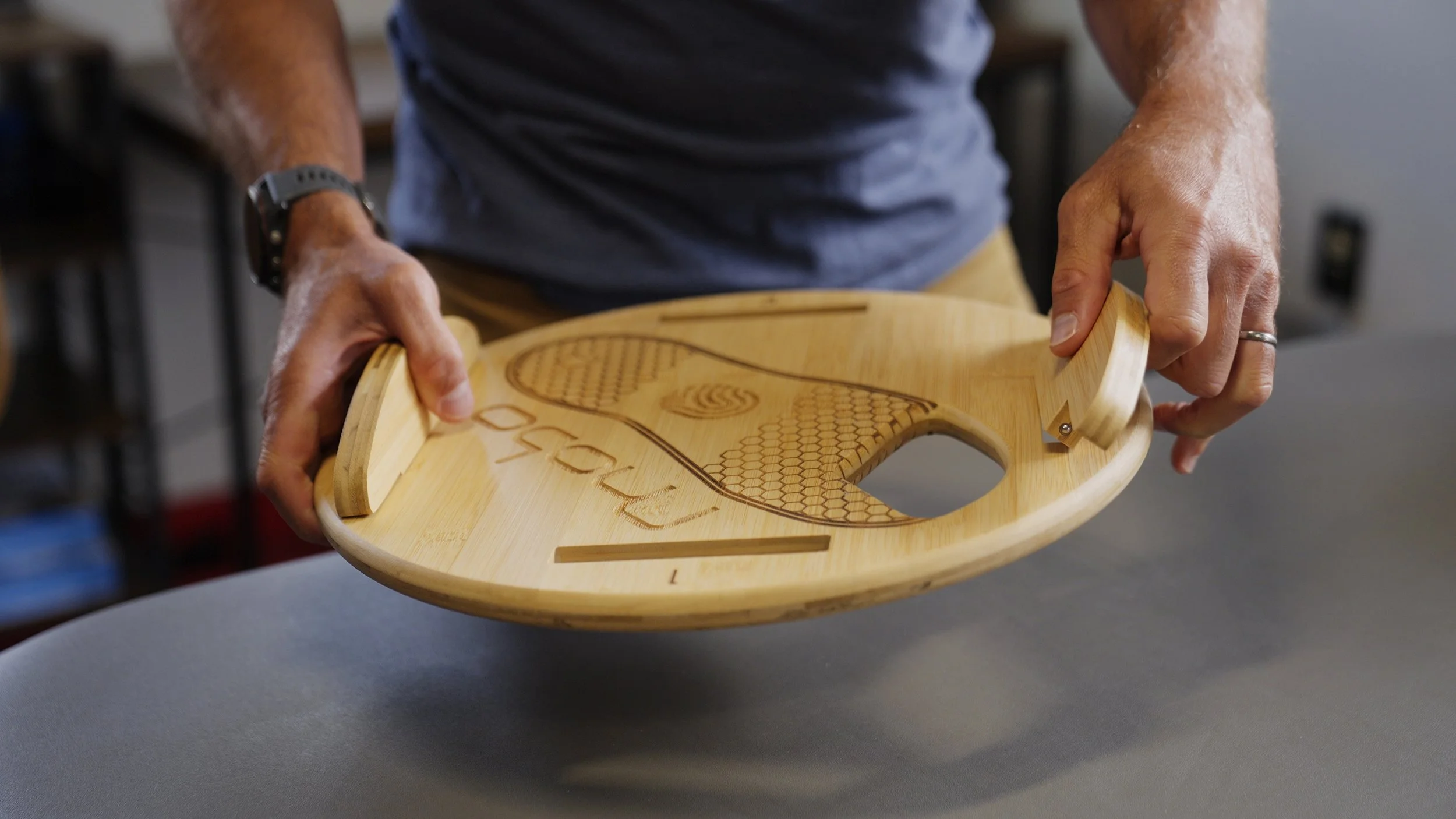 Person holding a round bamboo tray with engraved wood design and text, wearing a gray shirt and watch, standing at a gray table.