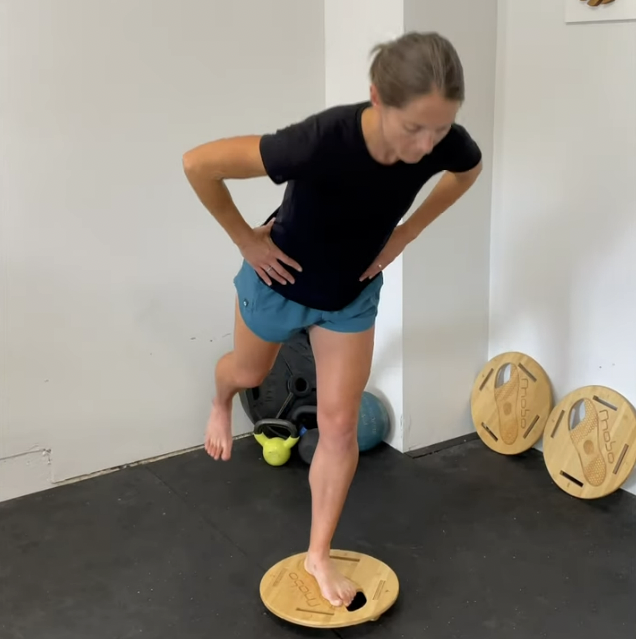 Woman doing a balance exercise on a wooden balance board in a gym.