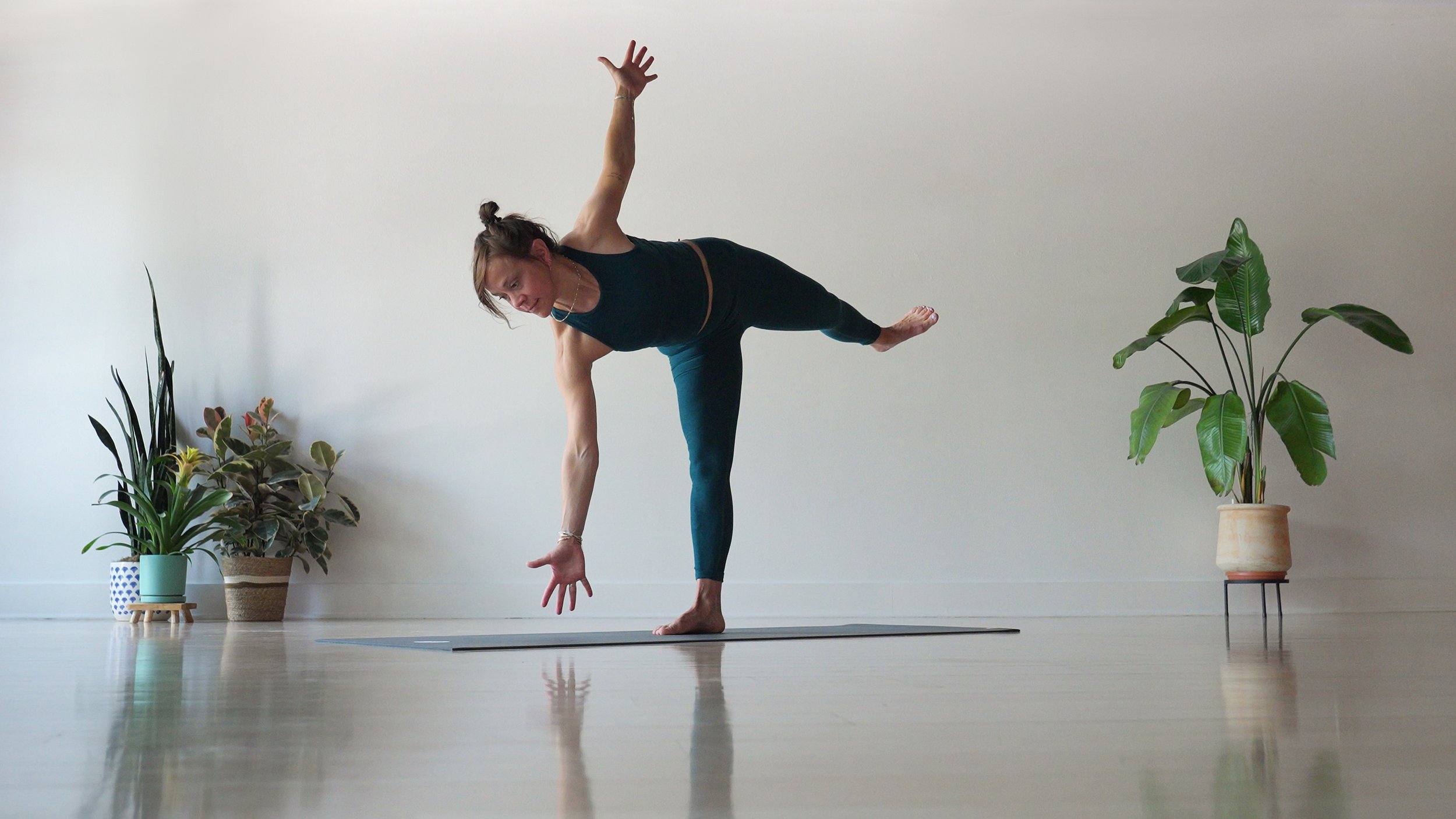 Woman practicing yoga indoors, balancing on one foot with her other leg extended to the side and her arm reaching downward, surrounded by potted plants on a polished wooden floor and plain wall background.