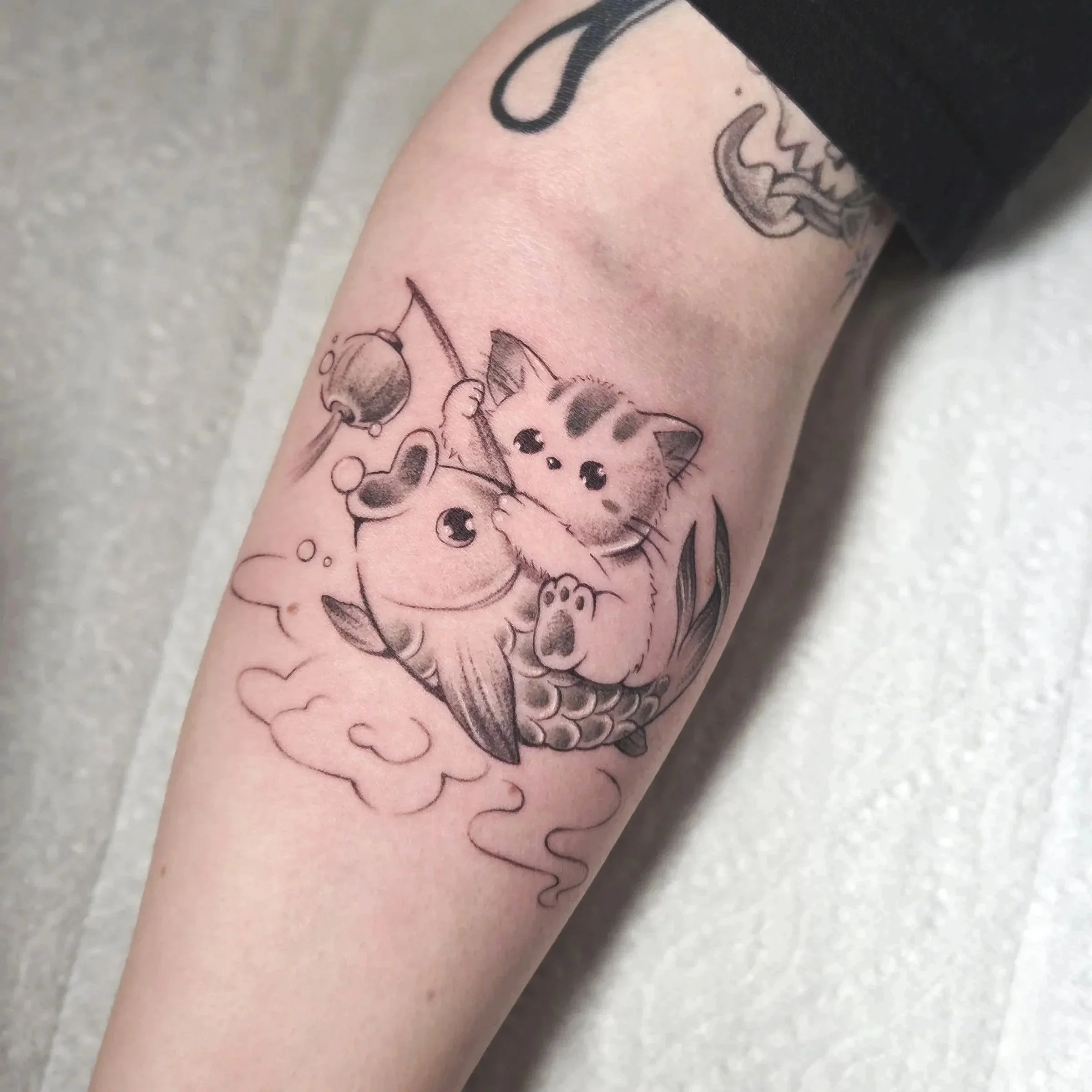Tattoo of a cute kitten holding a carp fish with a fishing hook, with clouds and bubbles in the background.