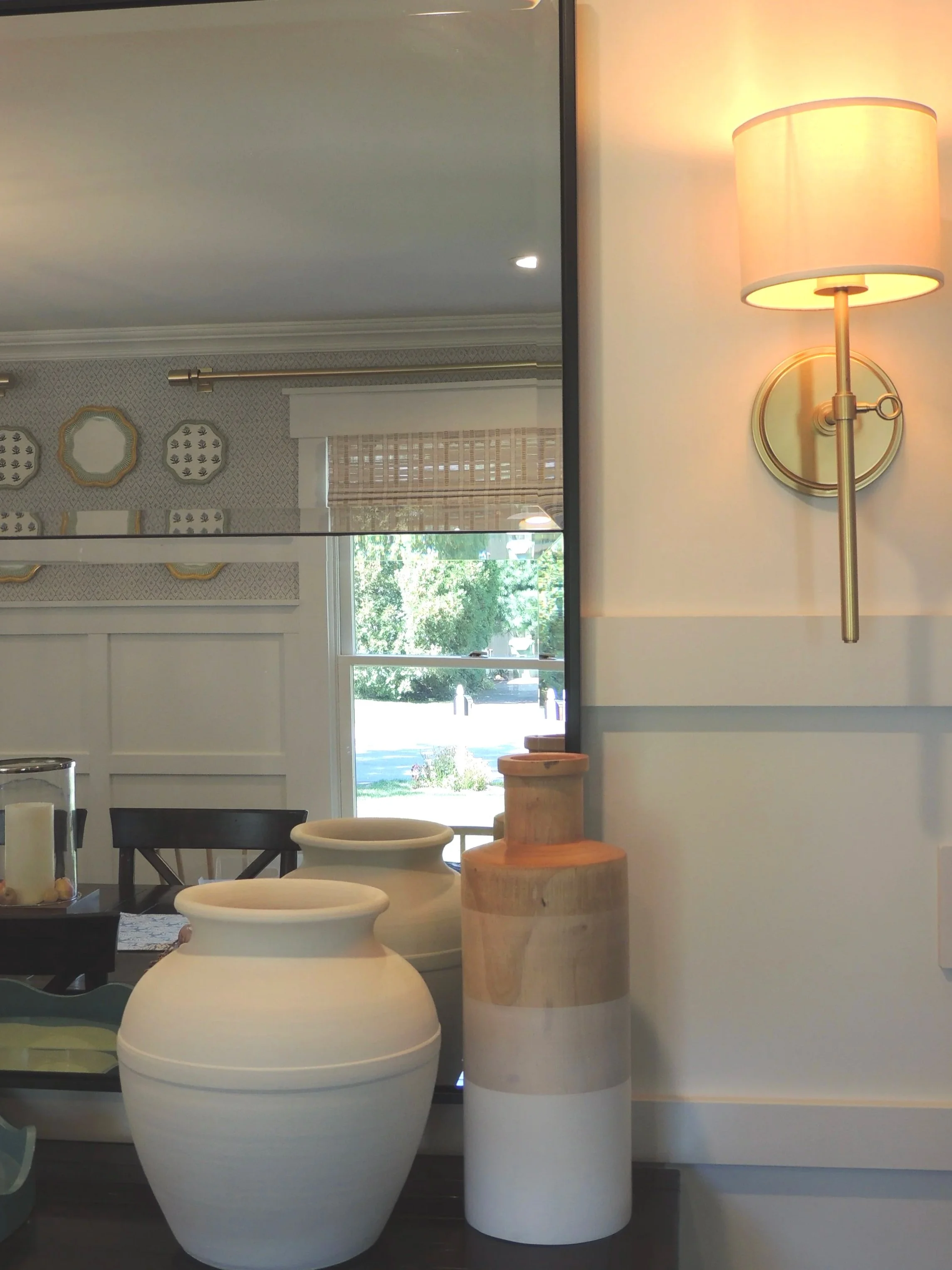 Interior view with large vases on a table, a wall with decorative plates, window with a view of greenery, and a wall-mounted lamp turned on.