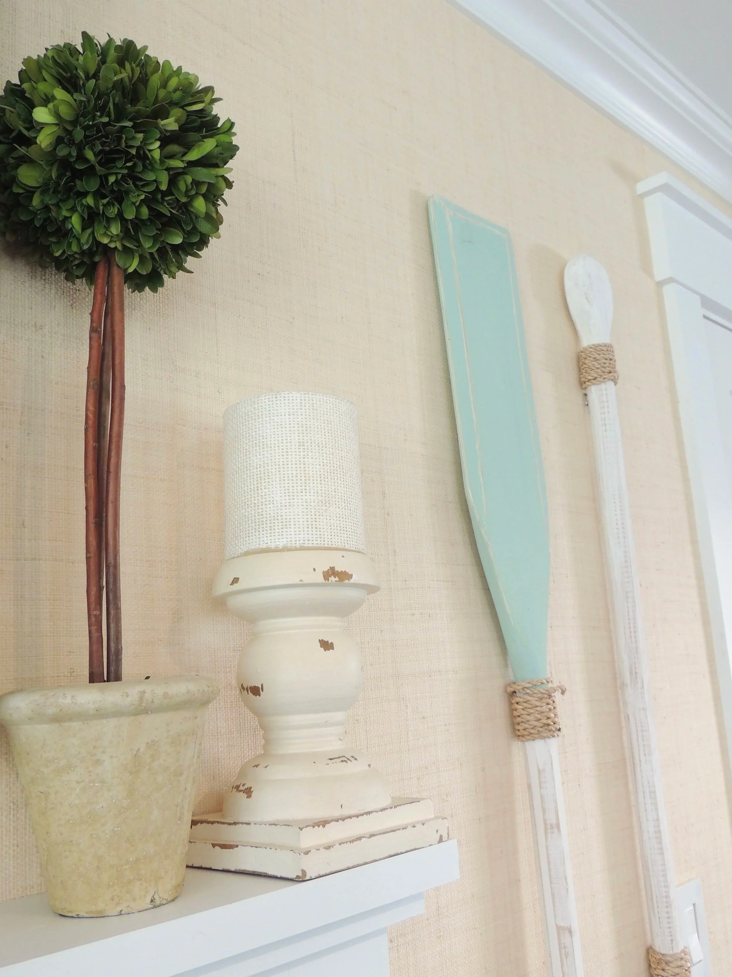 Decorative wall with potted topiary, distressed white table lamp with woven shade, light blue wooden paddle, and two white wooden oars with rope accents.
