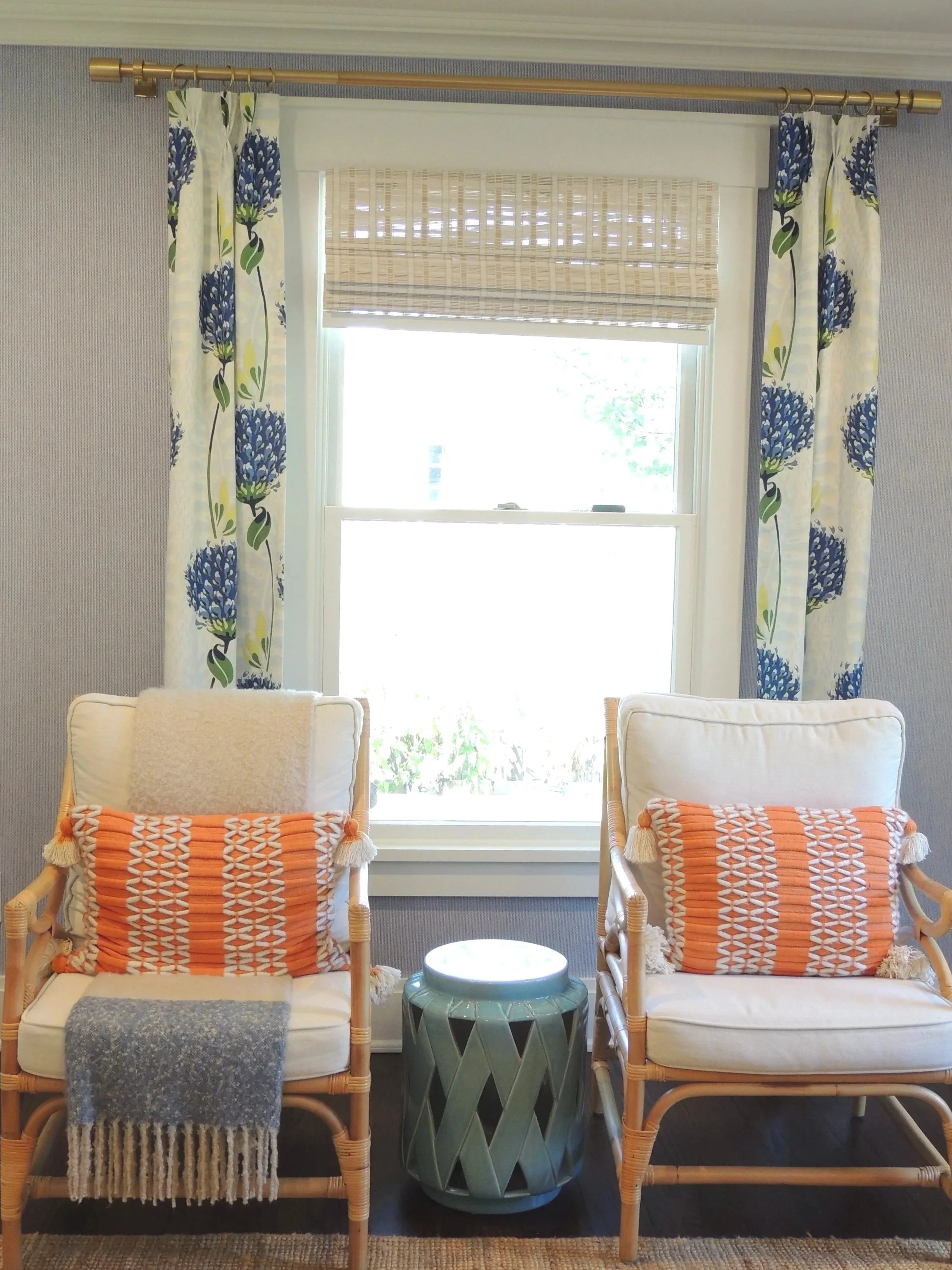 Living room window with blue and yellow floral curtains, bamboo chairs with orange patterned pillows, a small green side table, and a beige wall with a window shade.