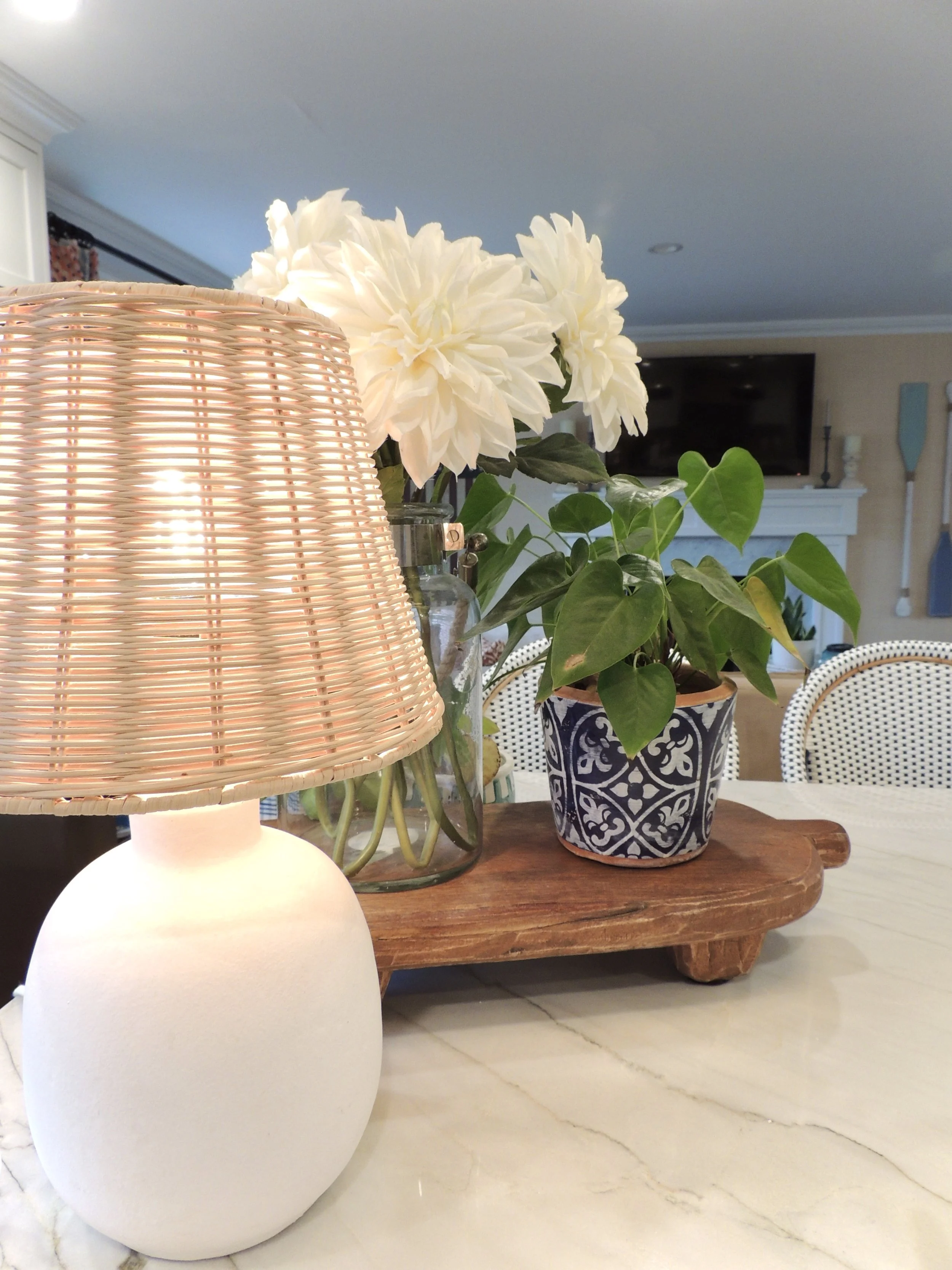 Table with a white lamp, a plant with white flowers and green leaves in a blue patterned pot, and a wooden tray, in a living room.