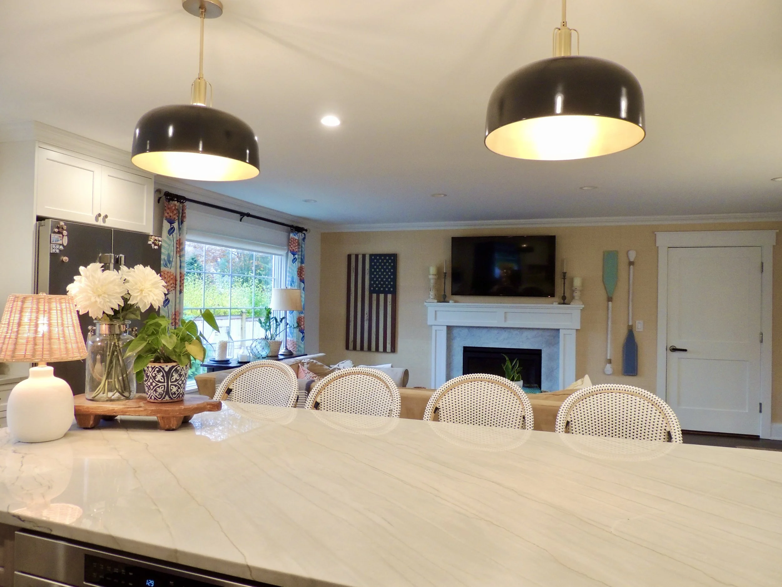 A home interior showing a kitchen and living room with black pendant lights, a large stone fireplace with a flat-screen TV above, a window with colorful curtains, and various decorative items.