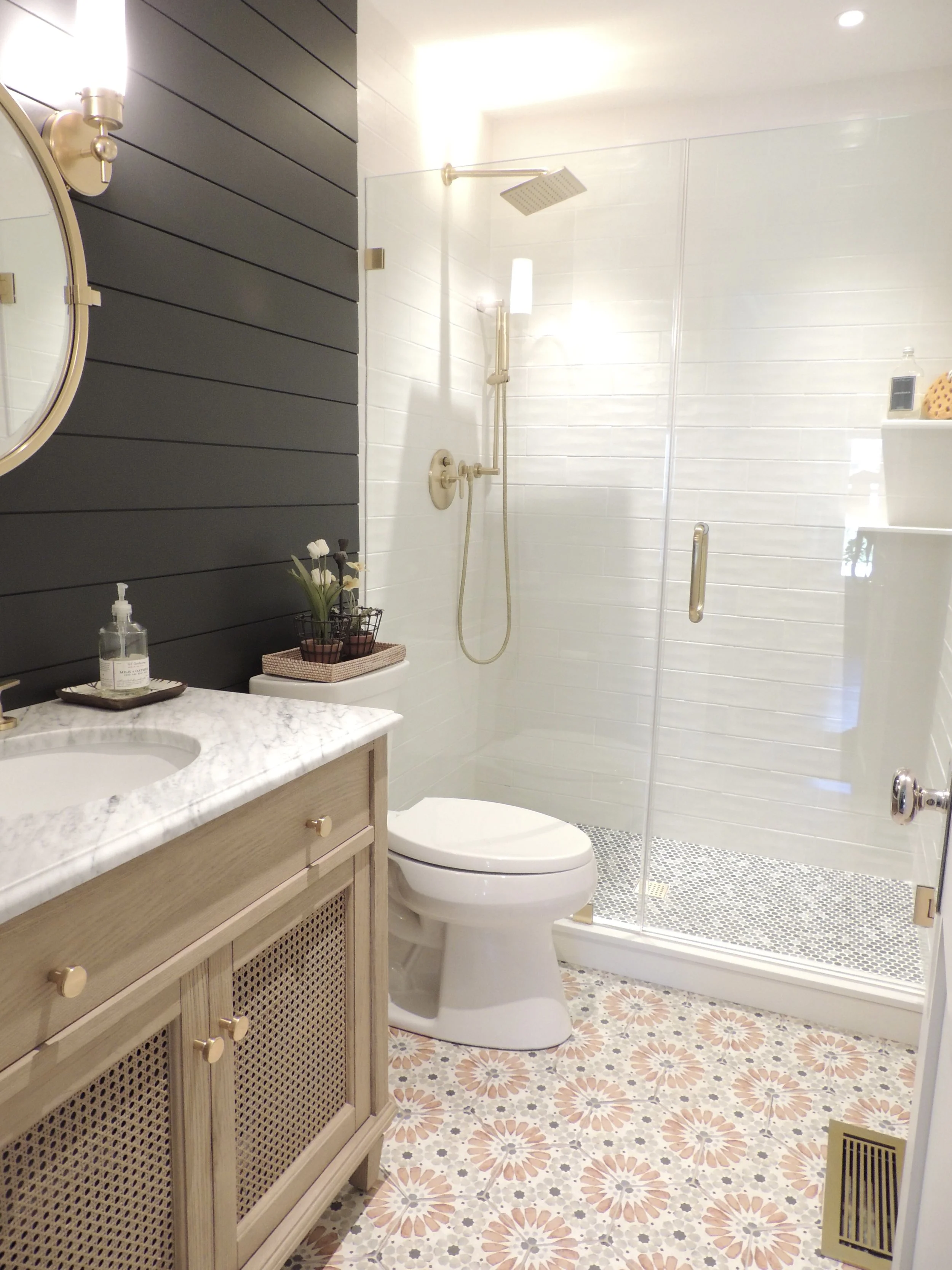 A modern bathroom with a marble vanity, toilet, and a walk-in shower with glass door. The sitting area has a patterned tile floor, a dark painted wall, and wall-mounted lights.