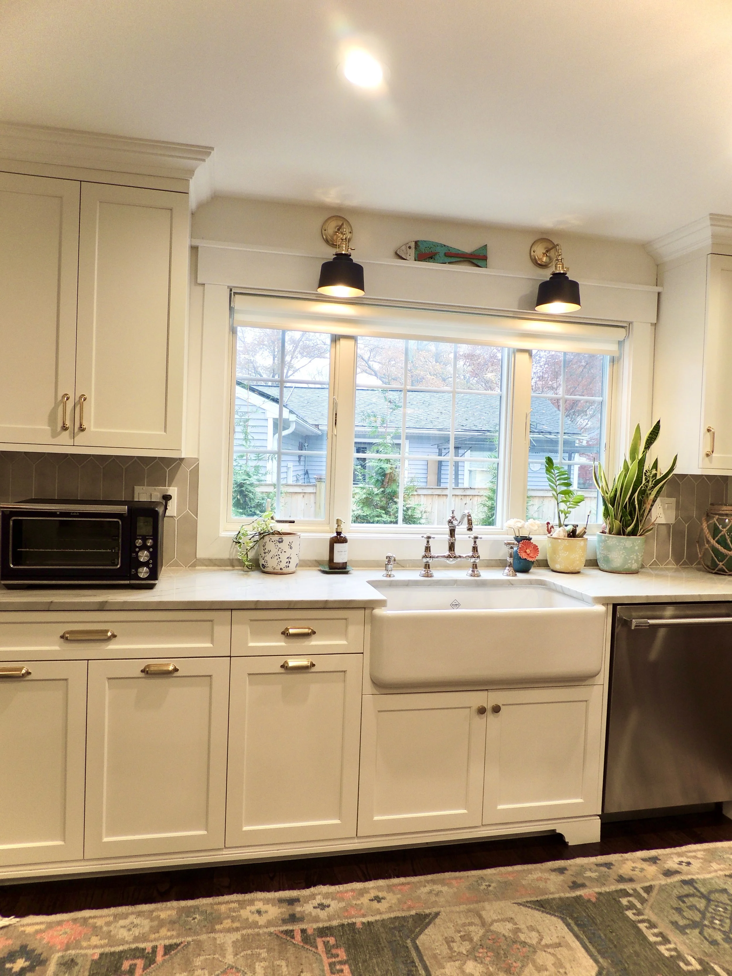 Kitchen with white cabinets and a farmhouse sink under a large window, decorated with potted plants, a soap dispenser, and a small cup of flowers. There is a toaster oven on the countertop and a colorful fish-shaped wall decoration above the window.