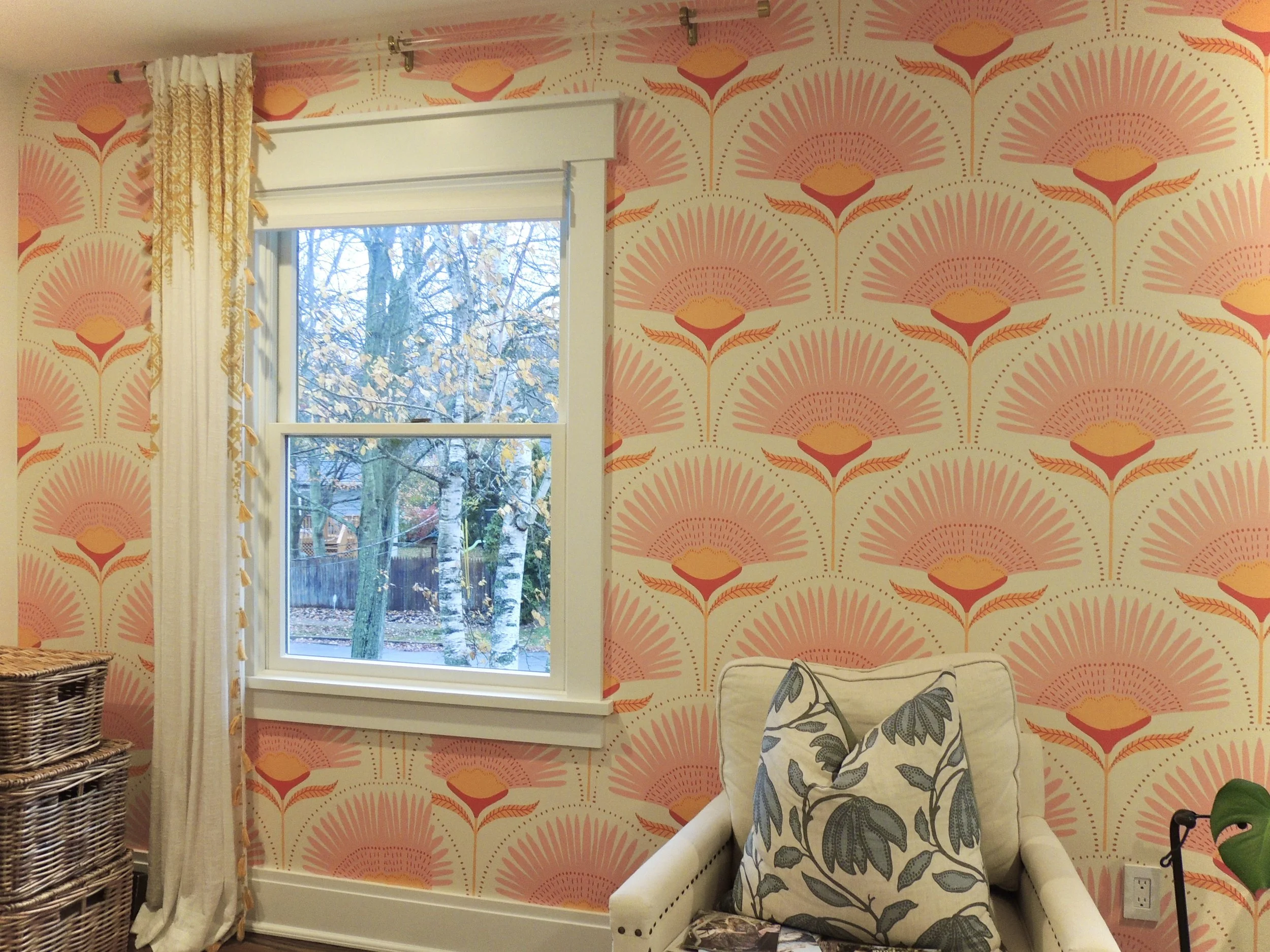 A room with a window showing a tree outside, pink and orange floral wallpaper, a cream armchair with a patterned pillow, and woven baskets to the side.