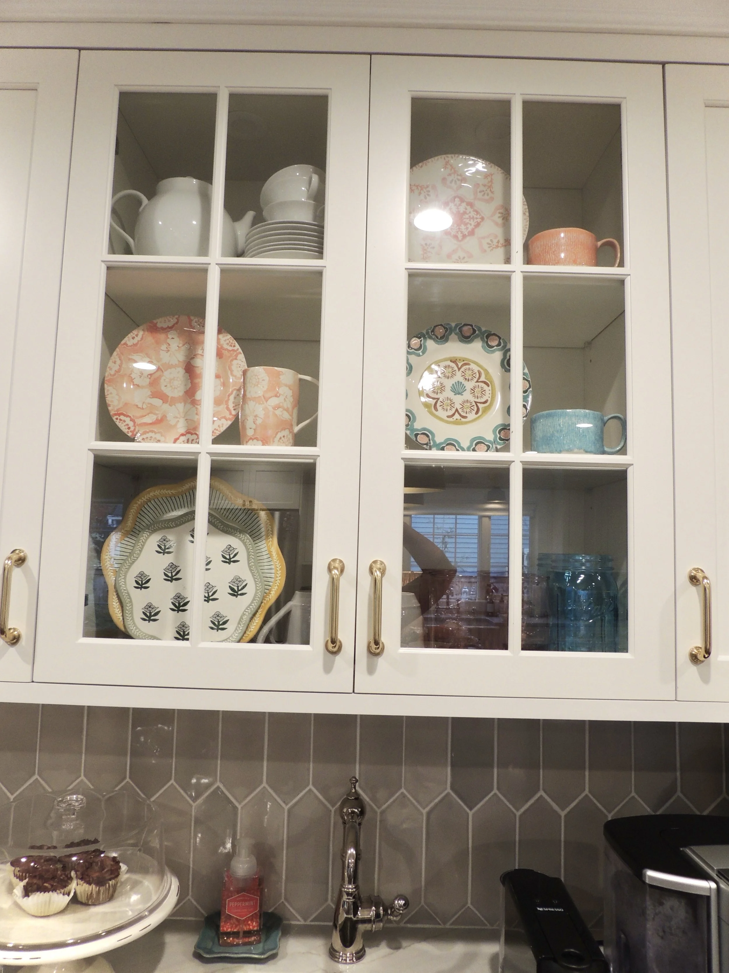 Kitchen cabinet with glass doors displaying dishes, cups, and bowls, with a counter below holding a cake, hand sanitizer, and kitchen appliances.