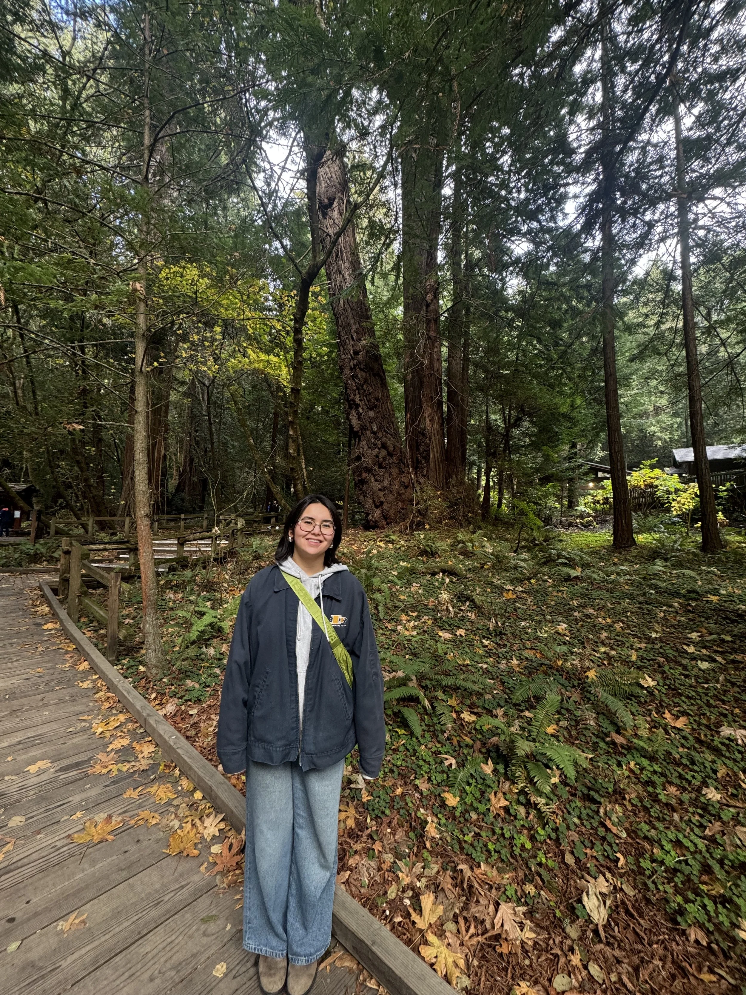 A young woman with glasses and dark hair standing on a wooden pathway in a forest with tall trees and fallen leaves.