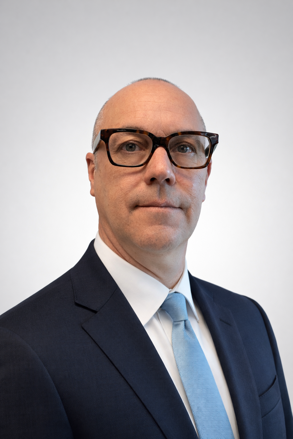 A man wearing glasses and a light blue collared shirt, smiling, indoors with blurred background.