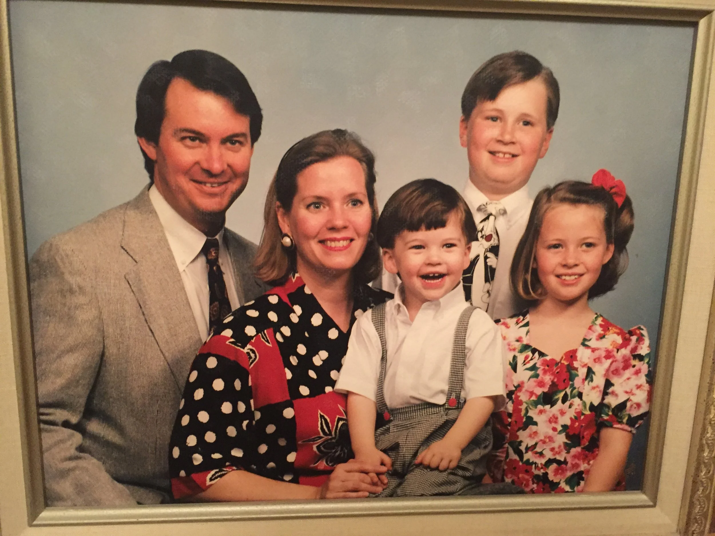 A family portrait of five people, two adults and three children, dressed in formal attire, posing against a light blue background.