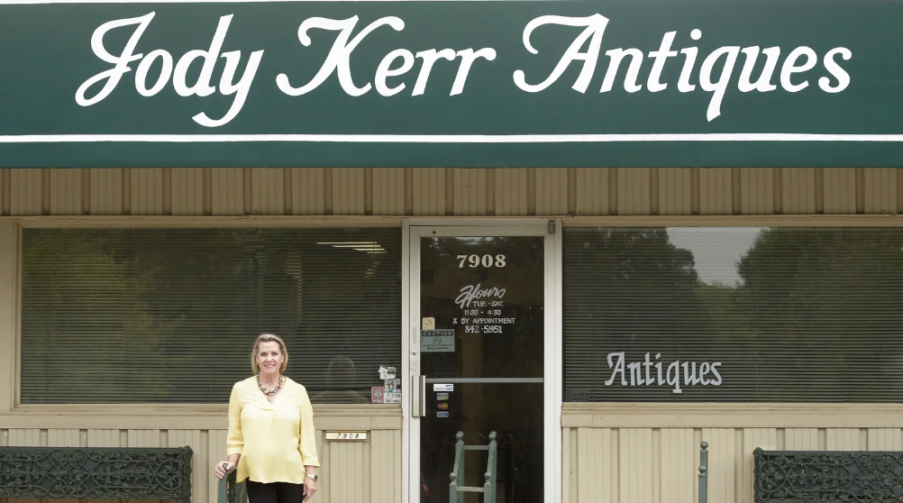 A woman standing in front of Jody Kerr Antiques store with a green sign and beige exterior, displaying the store's name and window signage that reads 'Antiques'.