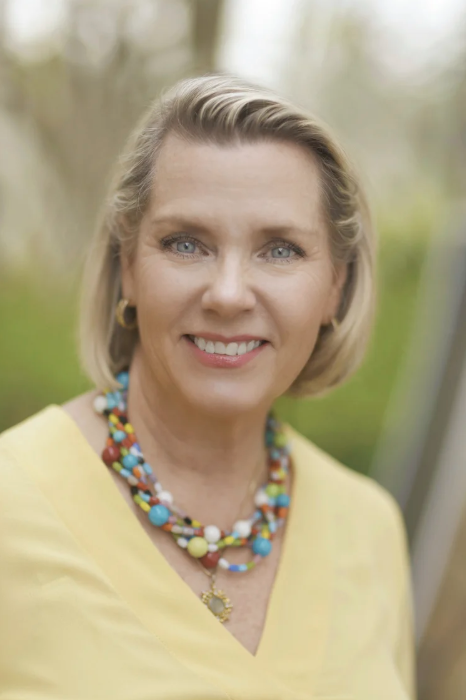 A middle-aged woman with blonde hair, blue eyes, and a bright smile, wearing a yellow top and colorful beaded necklaces outdoors.