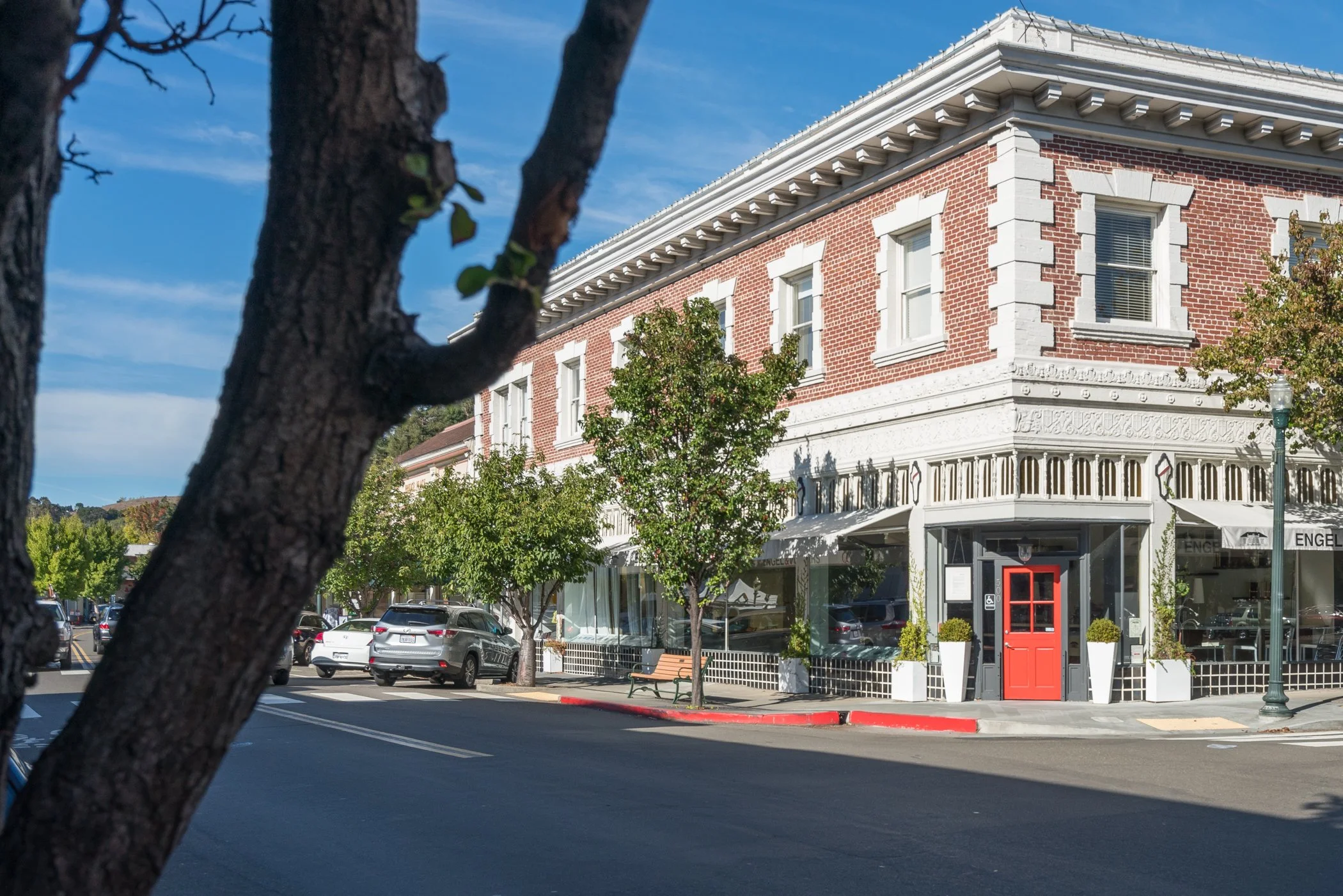 A street corner with a historic brick building featuring white decorative trim, a bright red door, trees, parked cars, a lamppost, and a clear blue sky.