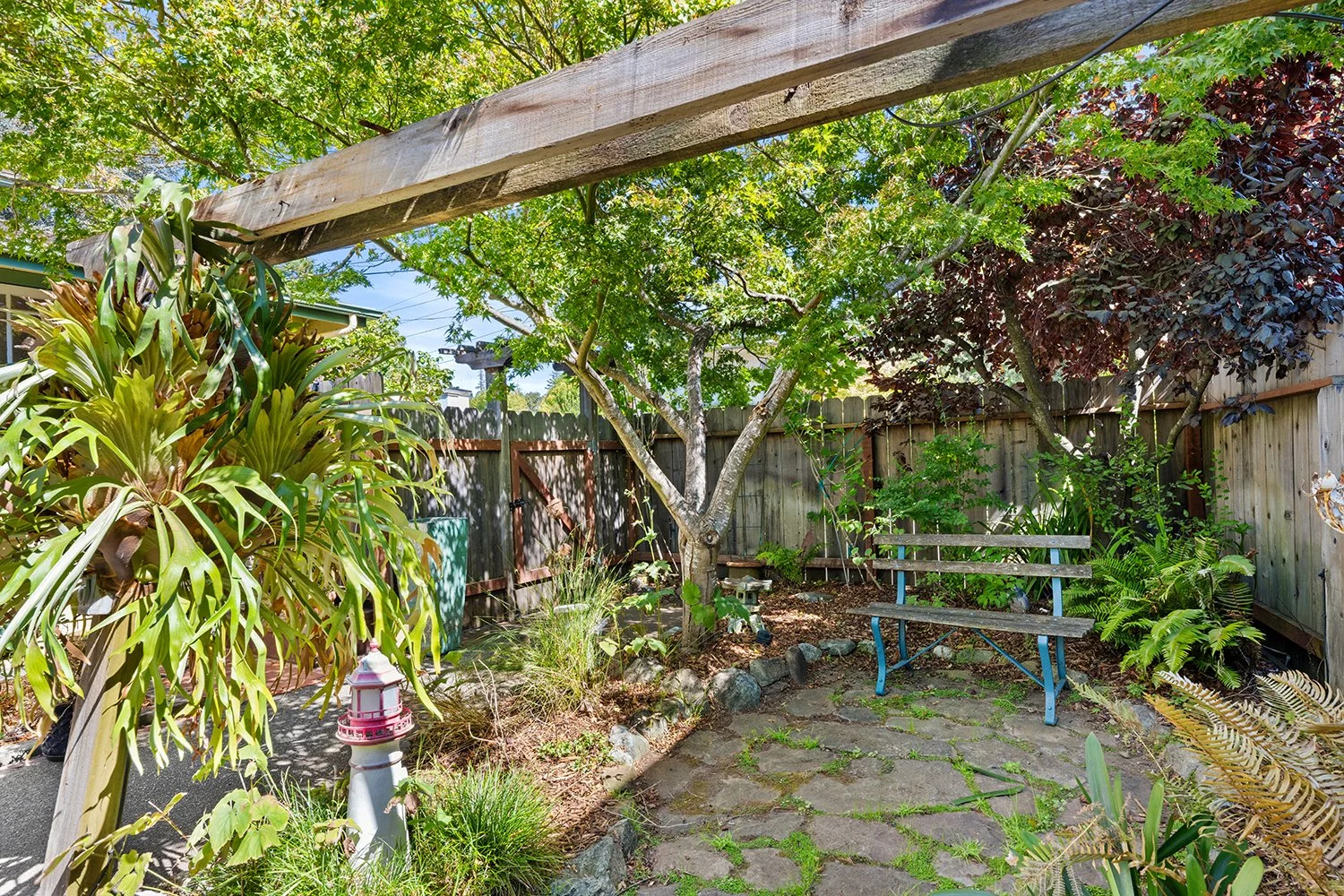 Backyard garden with trees, a wooden fence, a blue garden bench, plants, and a pink lanterned garden ornament.