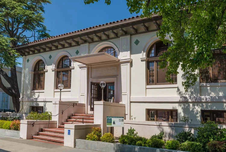 Front of a historic white building with arched windows and steps leading to an entrance, surrounded by green trees and landscaping.