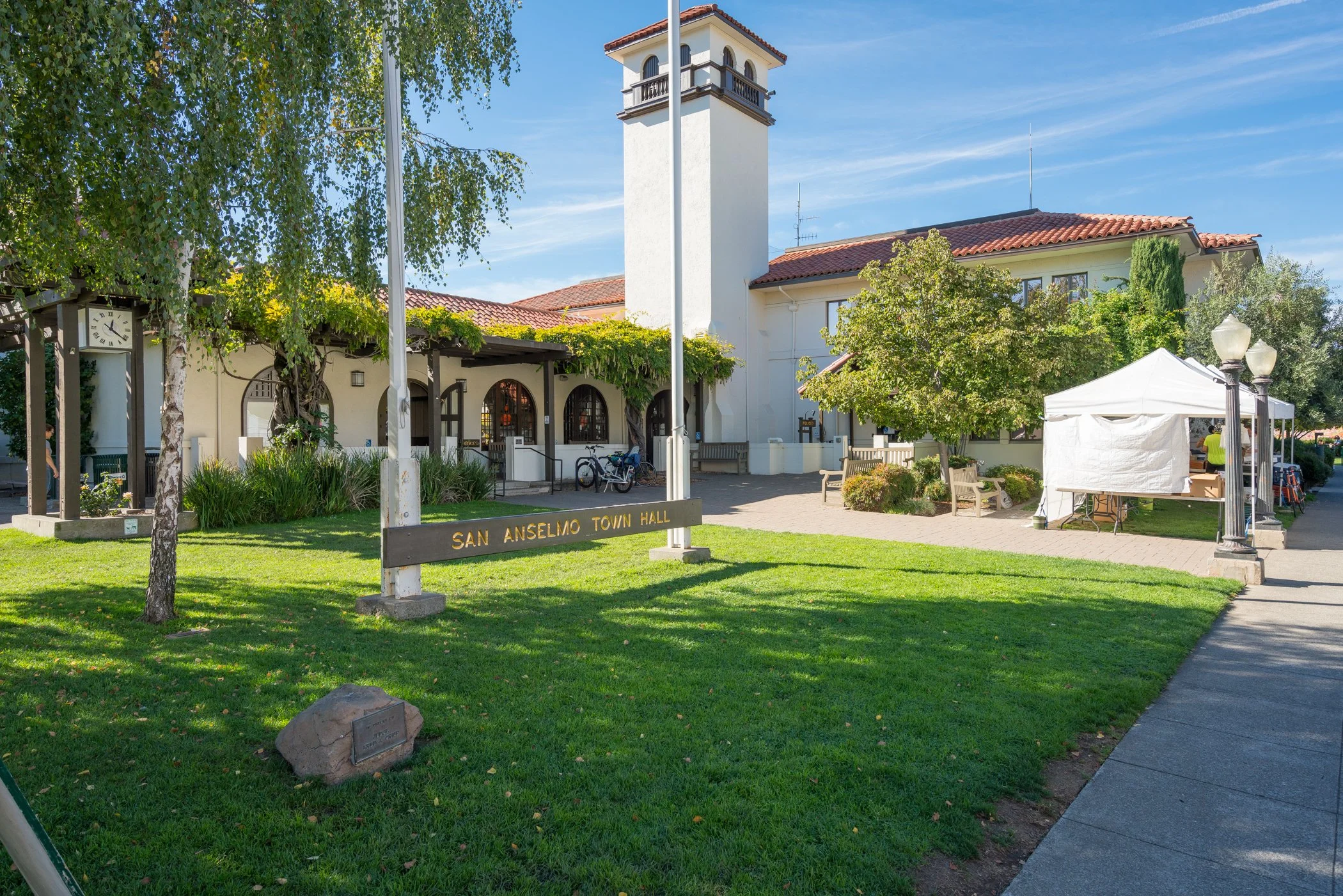 San Anselmo Town Hall exterior with a green lawn, trees, a white tent, benches, and a clock tower under a clear blue sky.