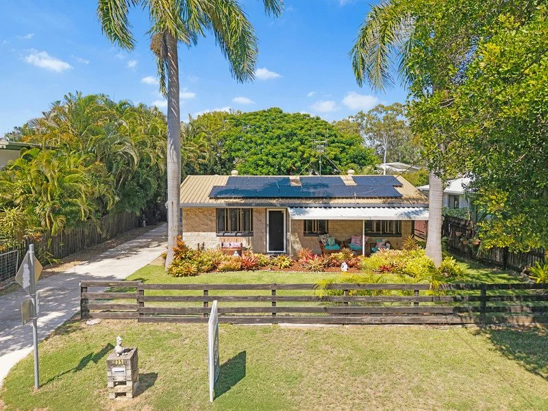 A single-story house with solar panels on the roof, surrounded by lush green trees and a garden with colorful plants. There is a fenced front yard with a mailbox and a driveway on the left side.