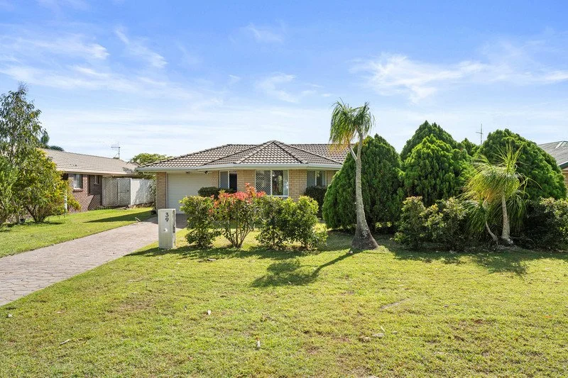 Single-story house with a tiled roof, surrounded by lush green yard and tropical plants, in a sunny suburban neighborhood.