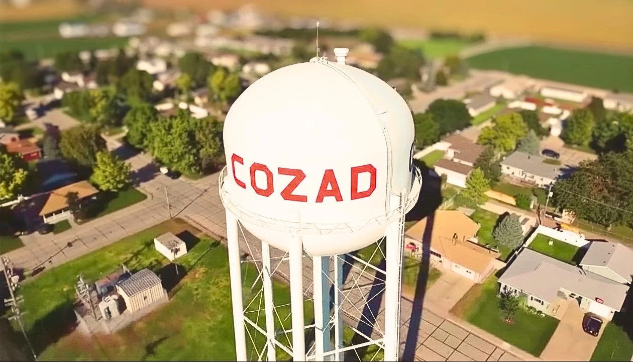 An aerial view of a water tower with the word 'COZAD' written on it in red letters, situated in a residential neighborhood with houses, trees, and streets.