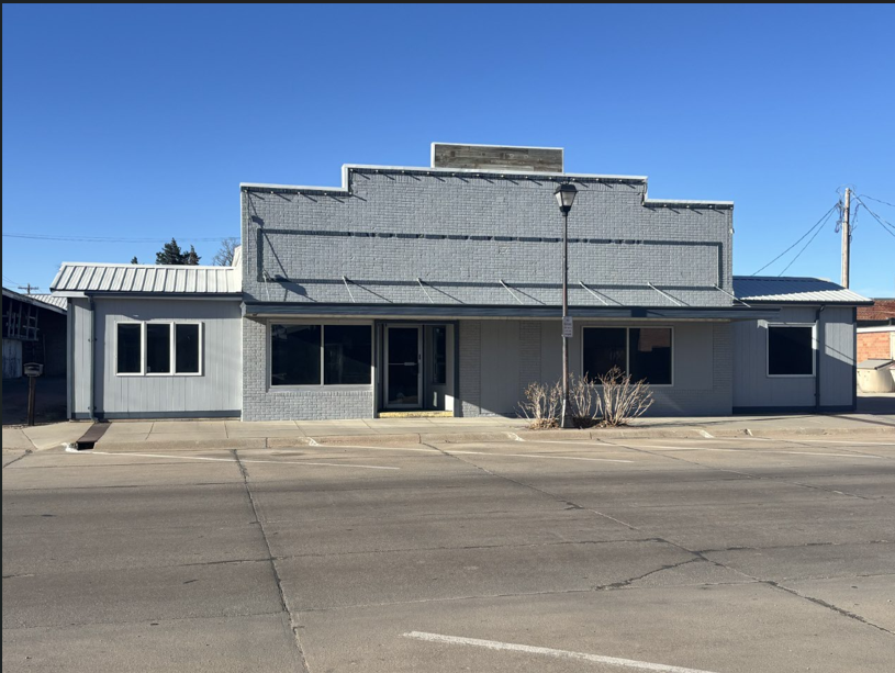 A small, single-story commercial building with a flat front facade, gray brick exterior, and large window panes, located on a street with a clear blue sky overhead.