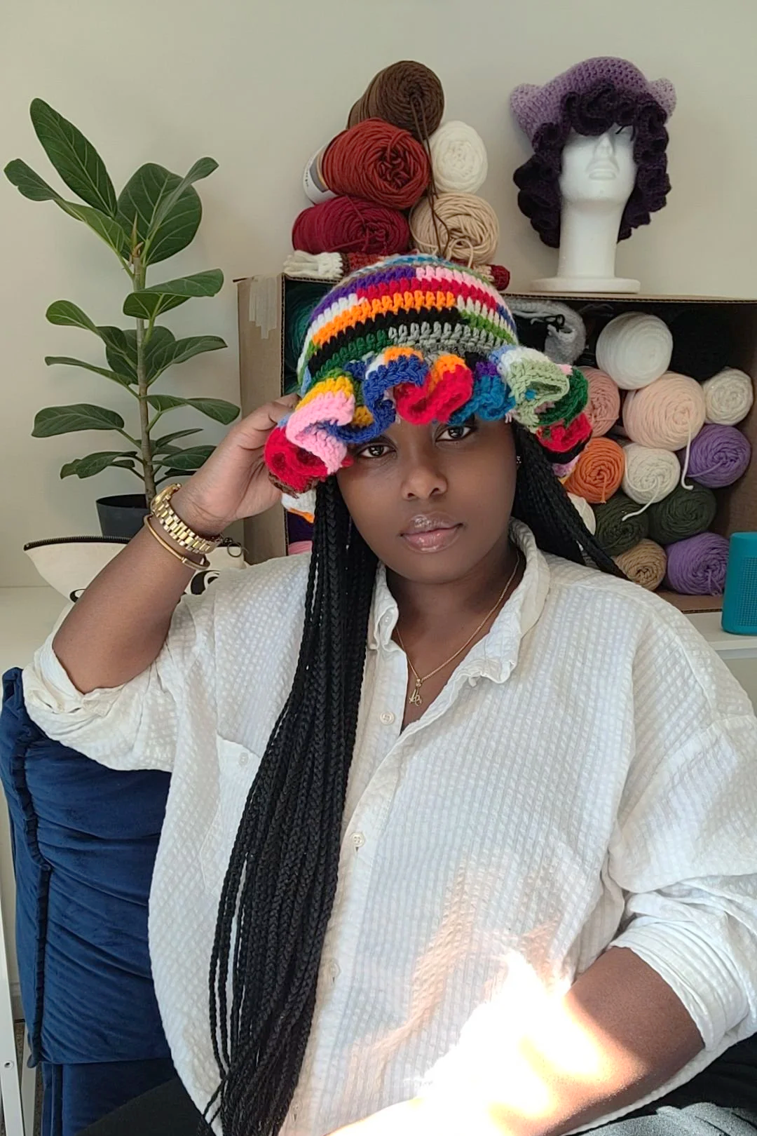 A woman with long black braids wearing a white shirt, holding a colorful crocheted hat on her head, sitting in front of a shelf with yarn and a mannequin head with a purple crocheted hat.