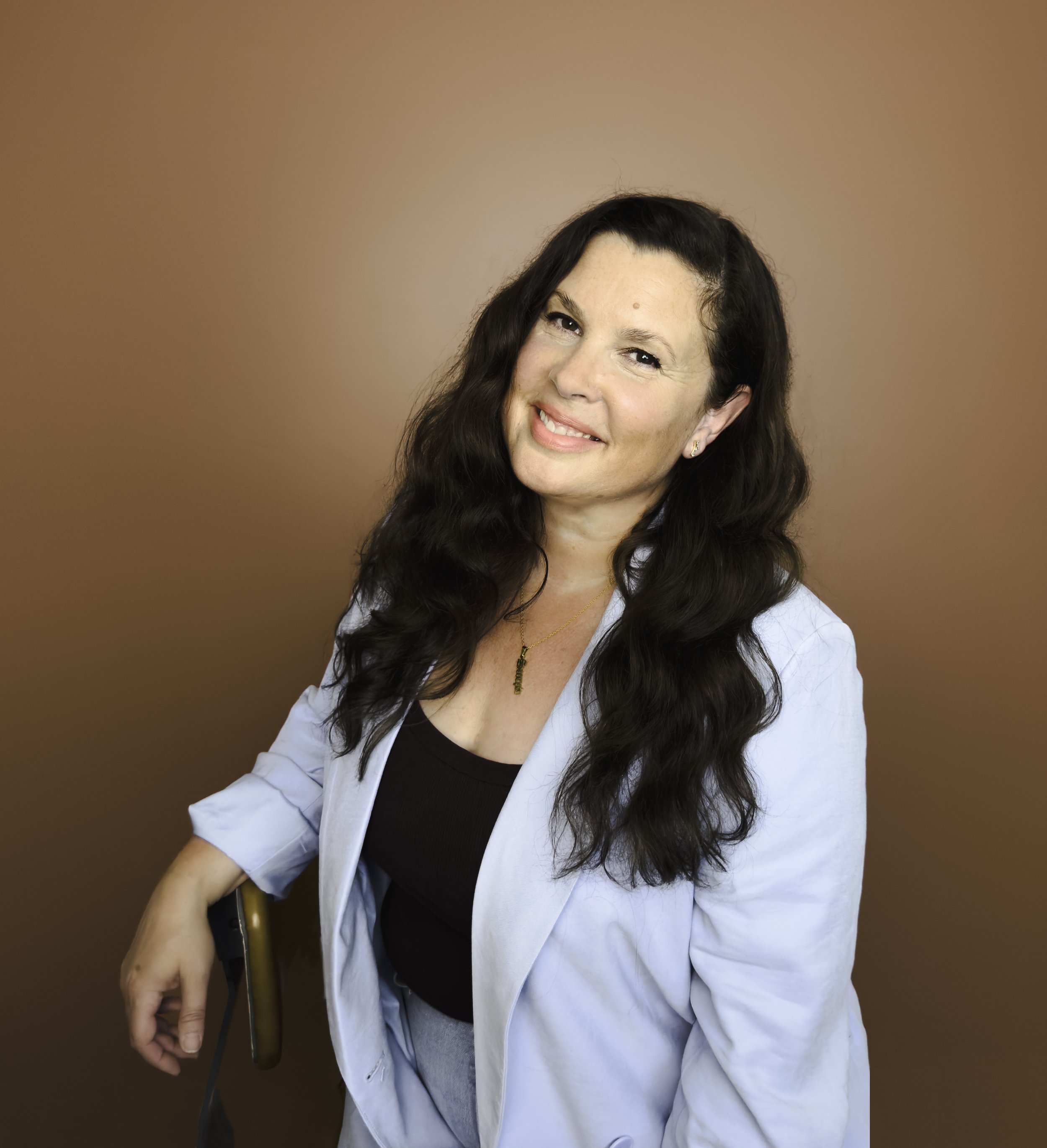 A woman with long, wavy dark hair, smiling, wearing a light blue blazer over a black top, with a gold necklace and earrings, standing against a brown background.