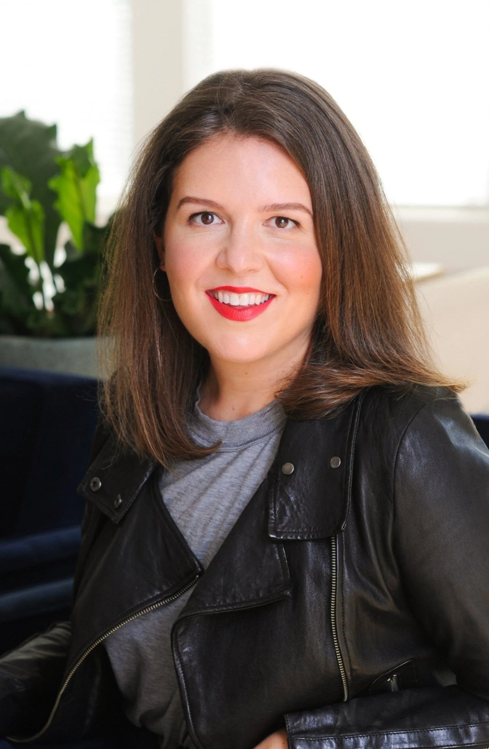 Portrait of a young woman with shoulder-length brown hair, red lipstick, gray t-shirt, and black leather jacket, smiling indoors with a plant and window in the background.