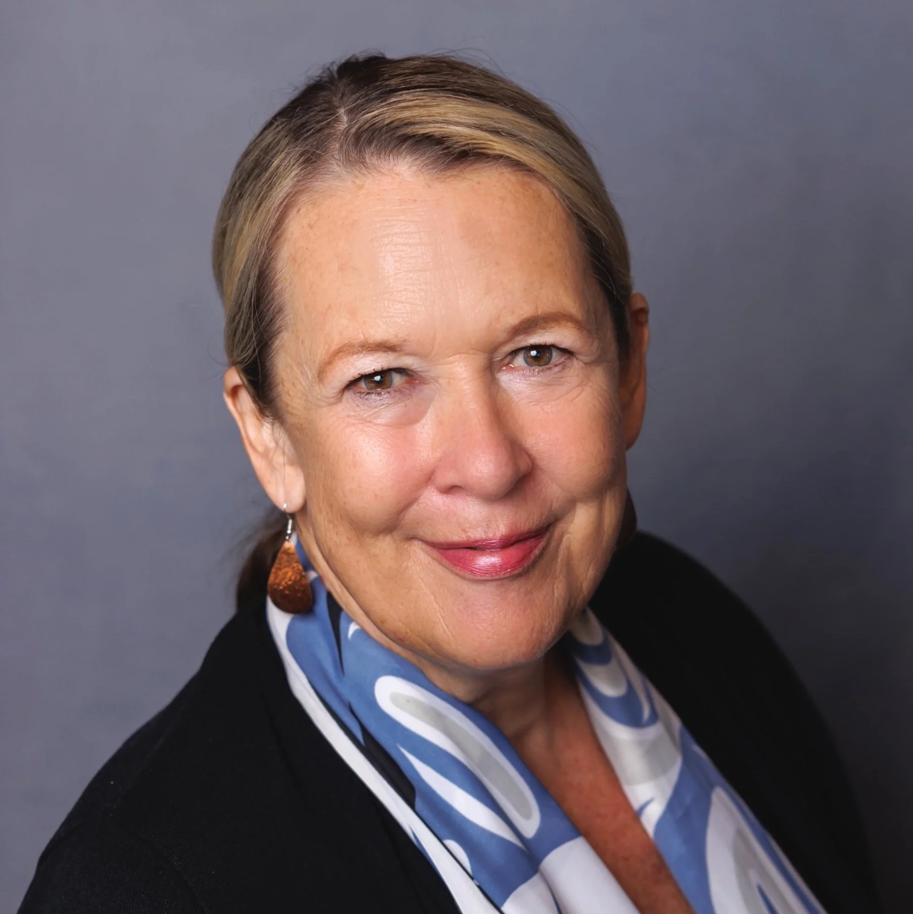 Professional headshot of a smiling middle-aged woman with blonde hair, wearing a black blazer and a blue patterned scarf, against a gray background.