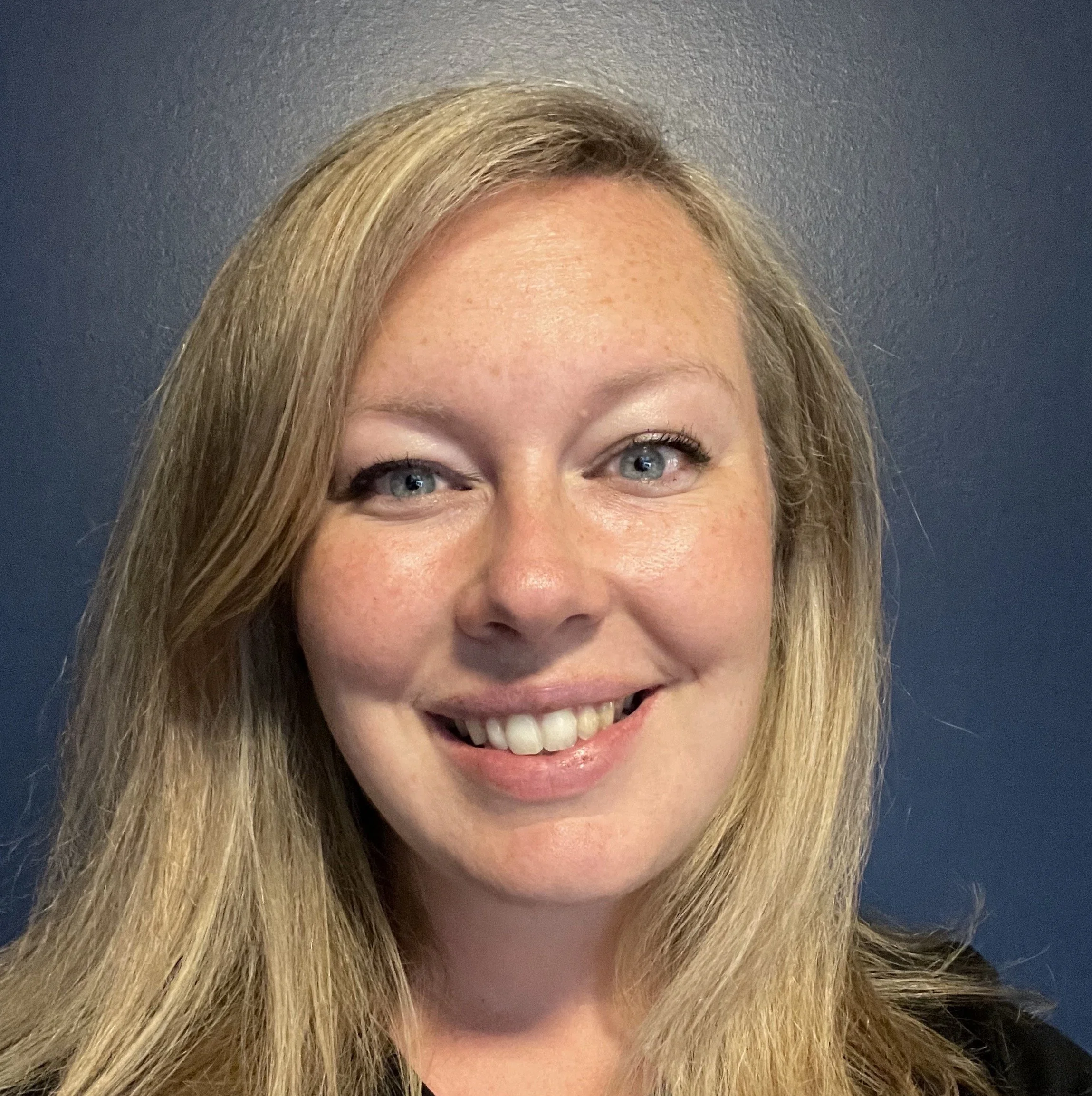 Close-up of a smiling woman with long blonde hair and blue eyes against a dark wall.