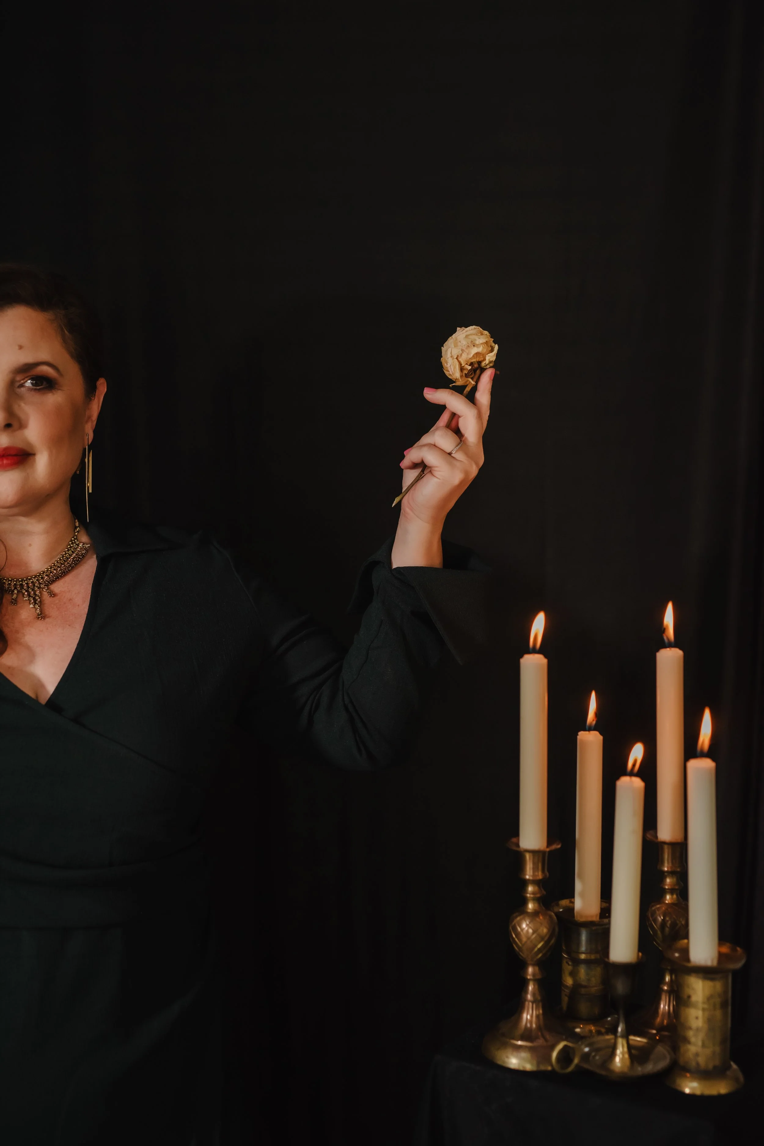 A woman with dark hair and gold jewelry, dressed in black, holding a dried flower in her right hand, standing beside a candelabrum with lit candles against a black background.