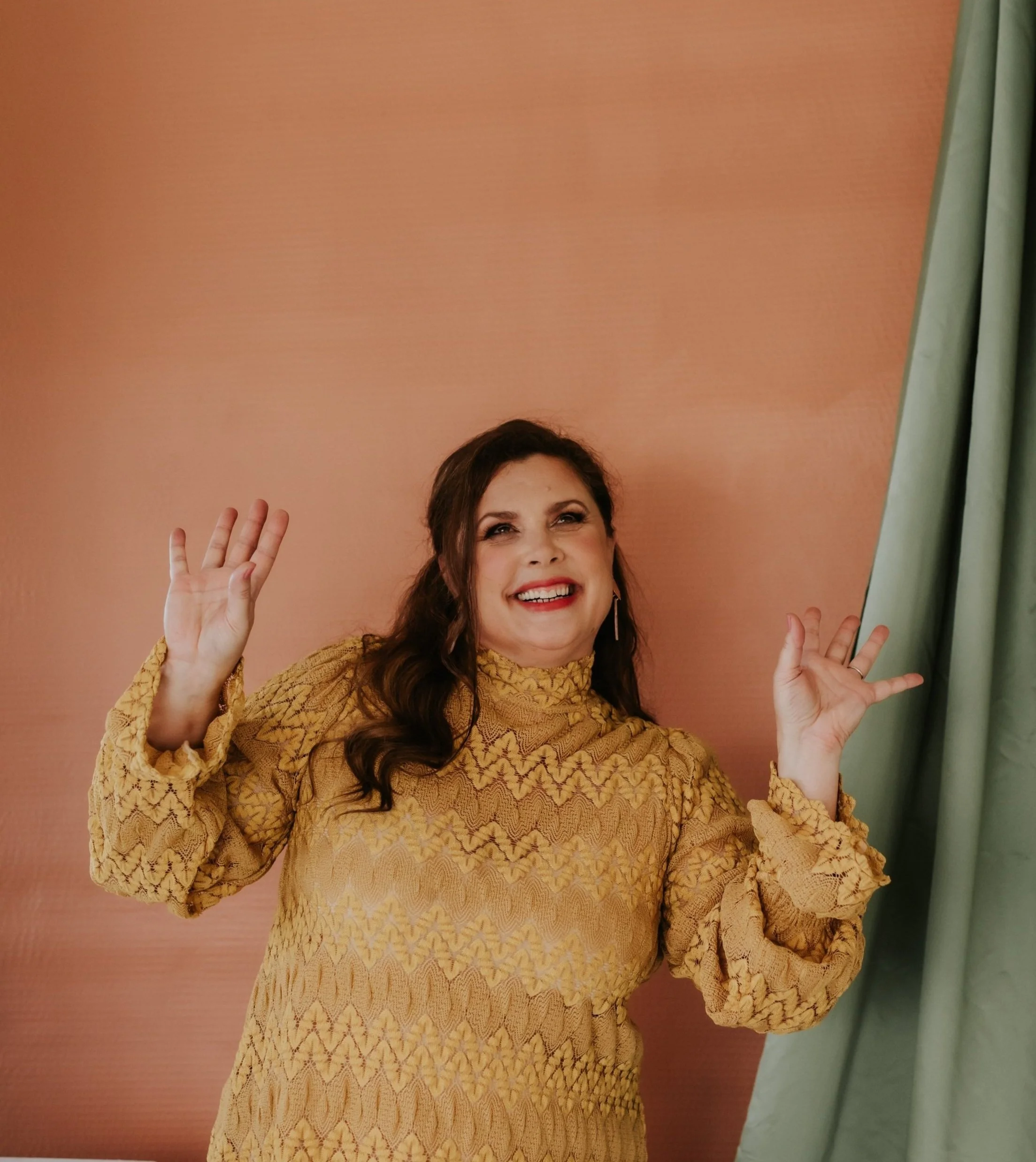 A woman with dark brown wavy hair smiling and waving, wearing a yellow patterned long-sleeve blouse, with a pink wall and green curtains in the background.