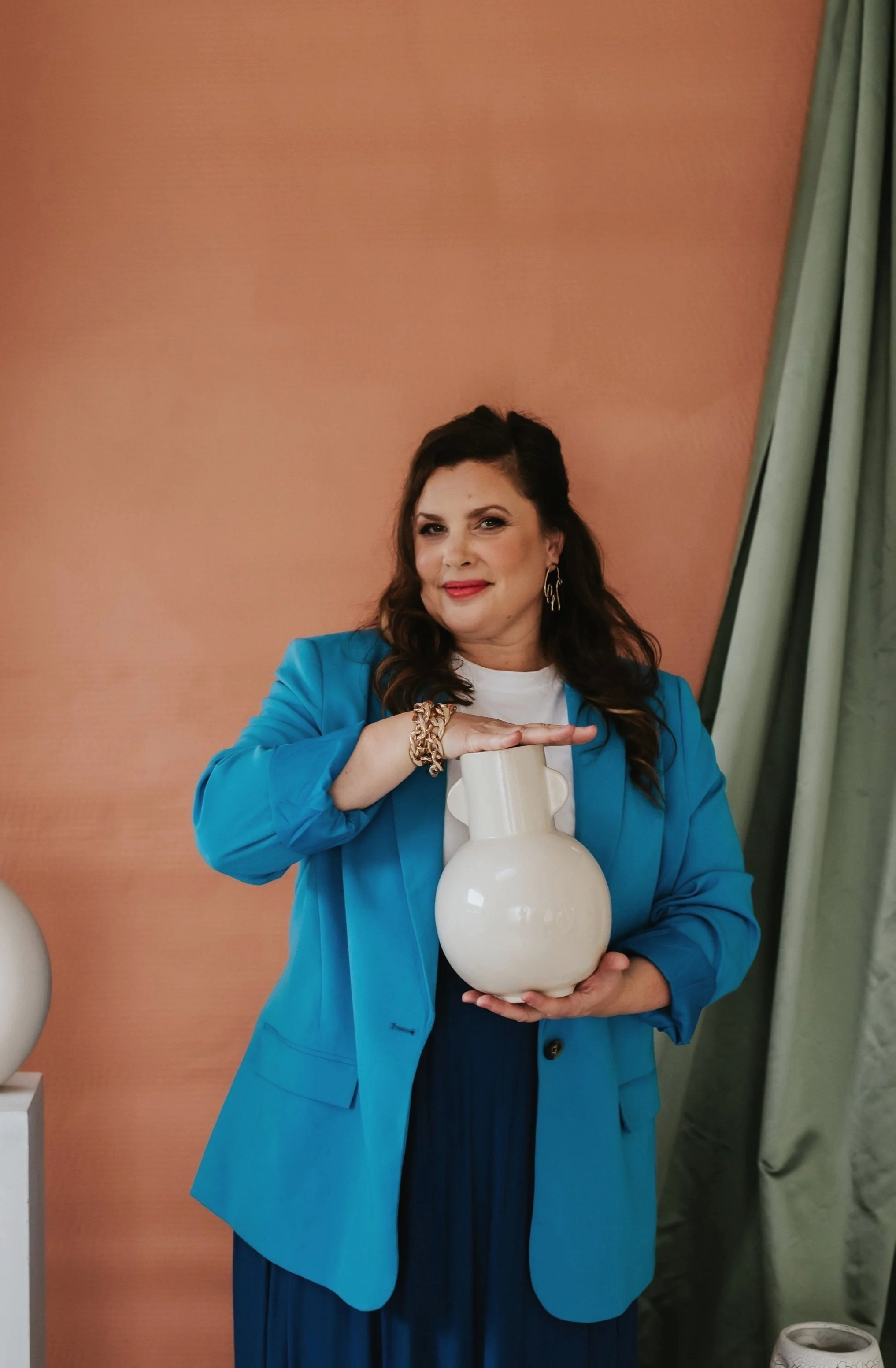 A woman with dark brown hair wearing a blue blazer, jewelry, and a white top, holding a large white vases while standing against a peach-colored wall with green curtains to her side.