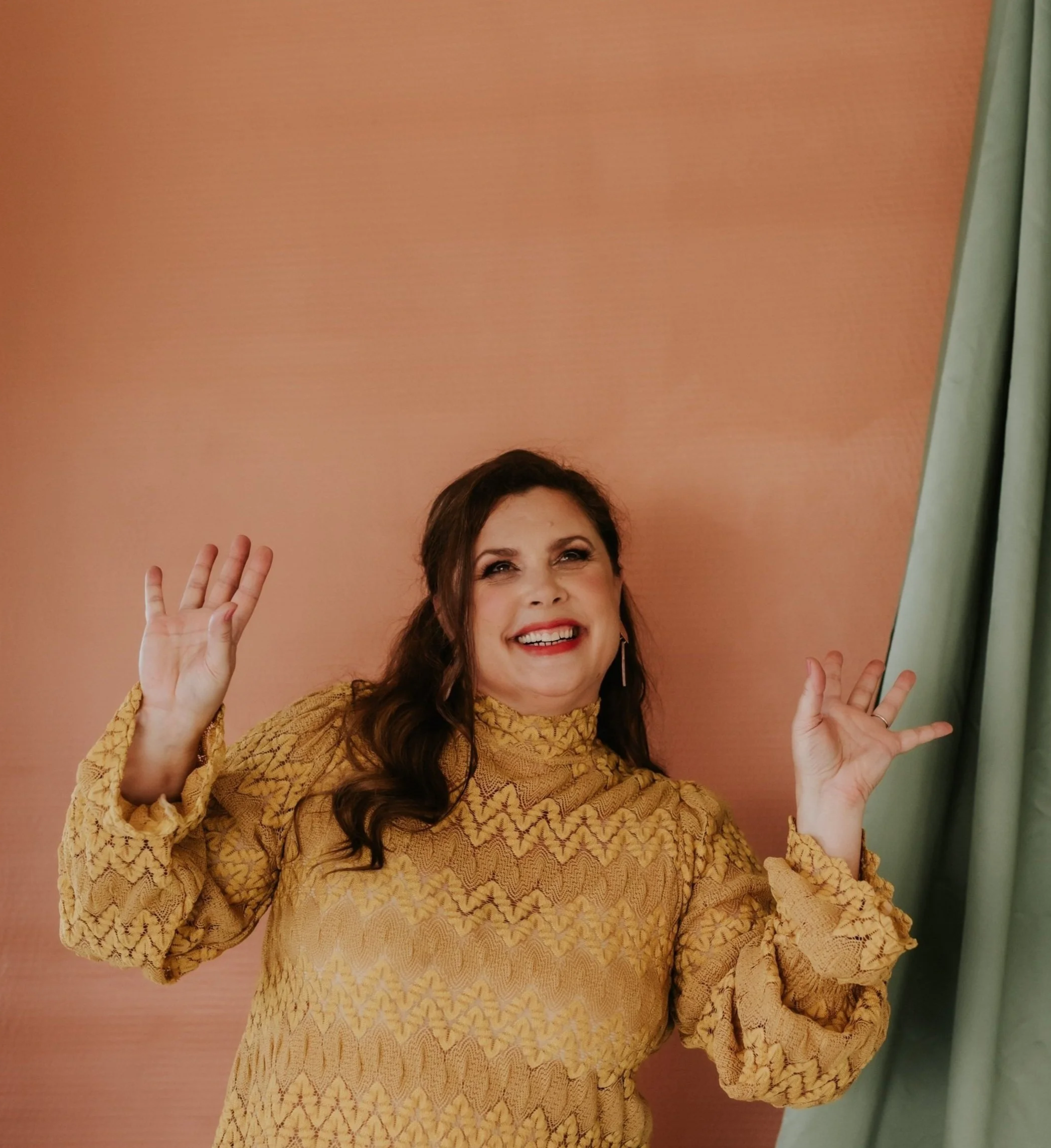 Woman with dark brown wavy hair smiling, wearing a yellow patterned high-neck blouse, posing with arms raised, against a pink background with a green curtain on the right.