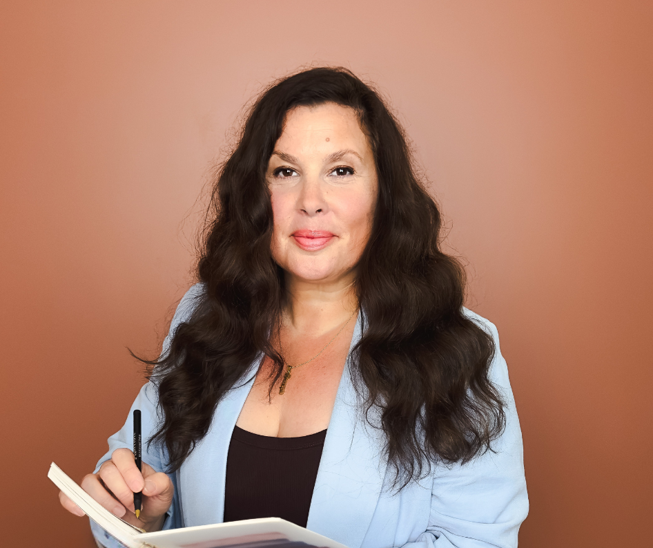 A woman with long, dark, wavy hair holding a pen and a notebook against a plain, warm-colored background.