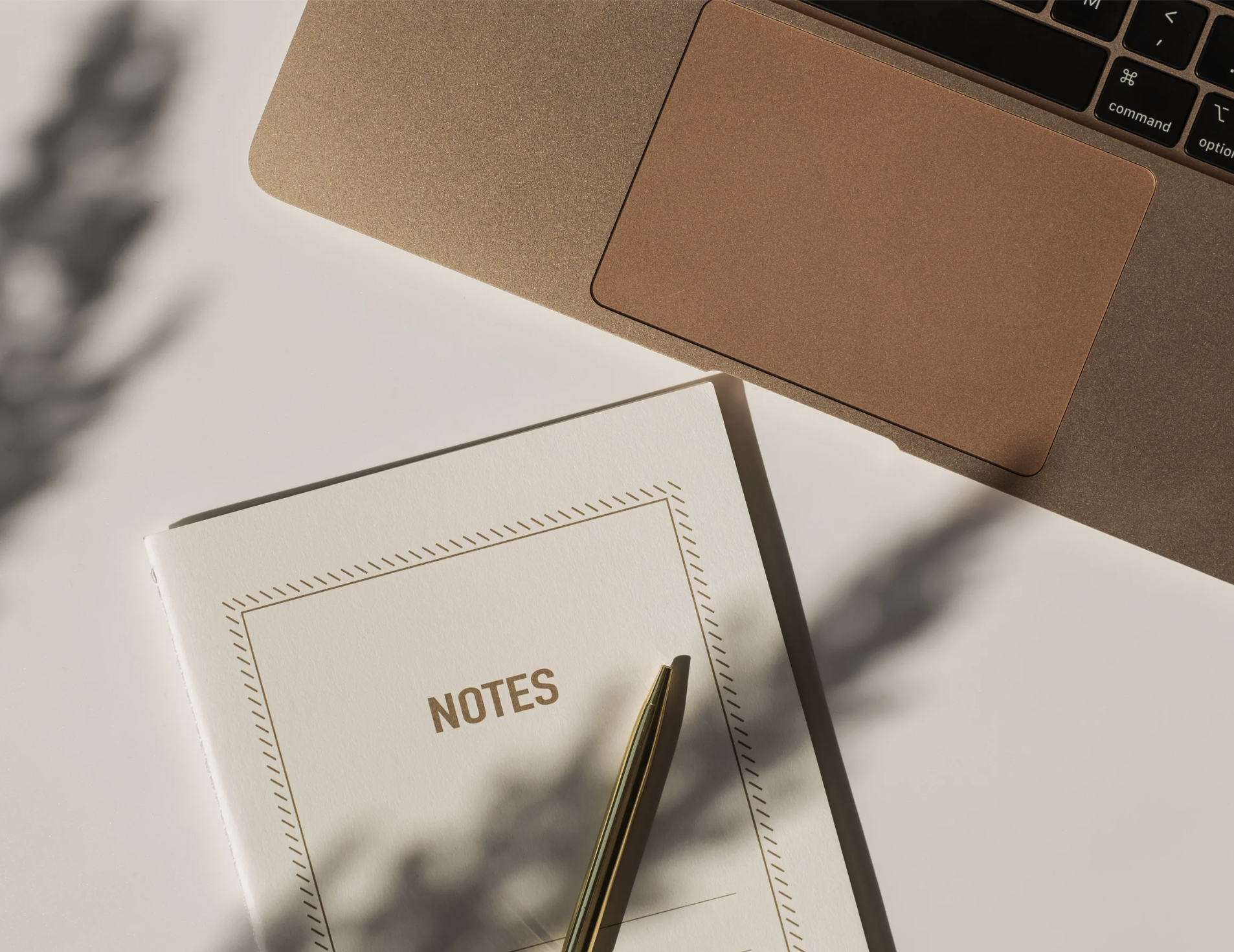 A close-up of a laptop keyboard, a notebook labeled 'NOTES' with a gold pen on top, and a shadow of a plant on a white surface.