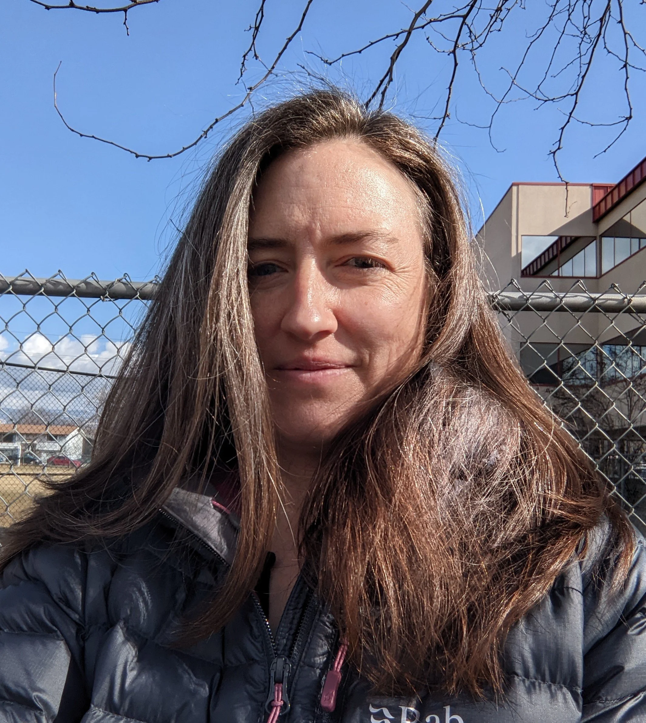 Headshot of a smiling middle-aged woman public speaking coaching and performance client  with brown hair, wearing a coat outdoors with blue sky in the background