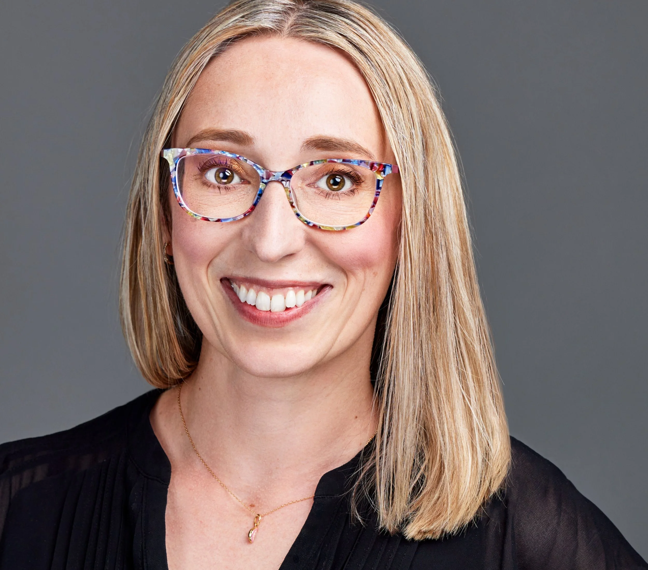 Portrait of a young woman public speaking coaching client with blond shoulder-length, black shirt, smiling indoors with a blue gray background