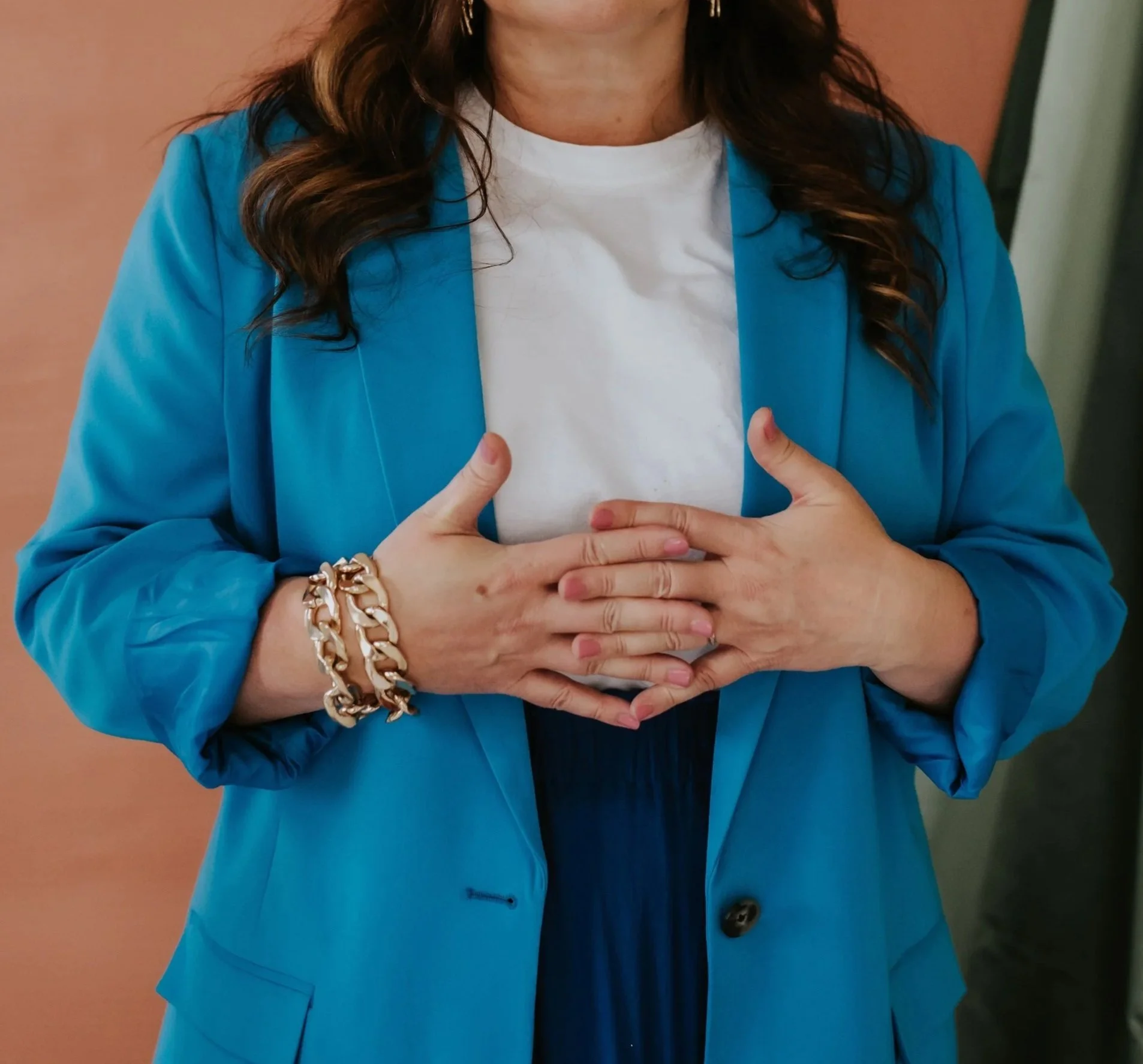 A woman wearing a blue blazer, white t-shirt, and dark pants, with her hands touching her upper stomach, wearing a chunky gold bracelet on her right wrist.